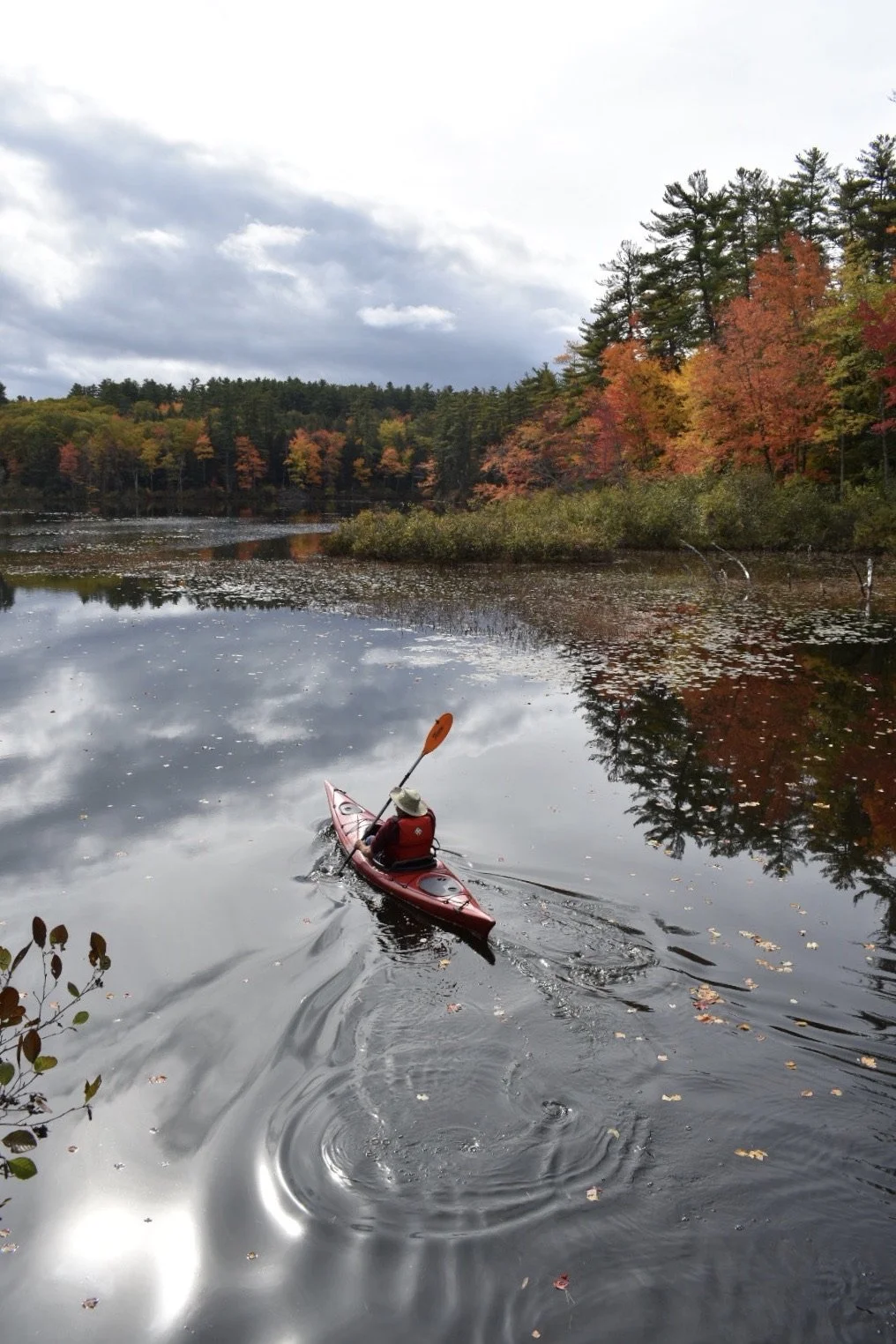 A person kayaking on a calm lake during fall with colorful orange, red, and green trees, and cloudy sky reflected in the water.