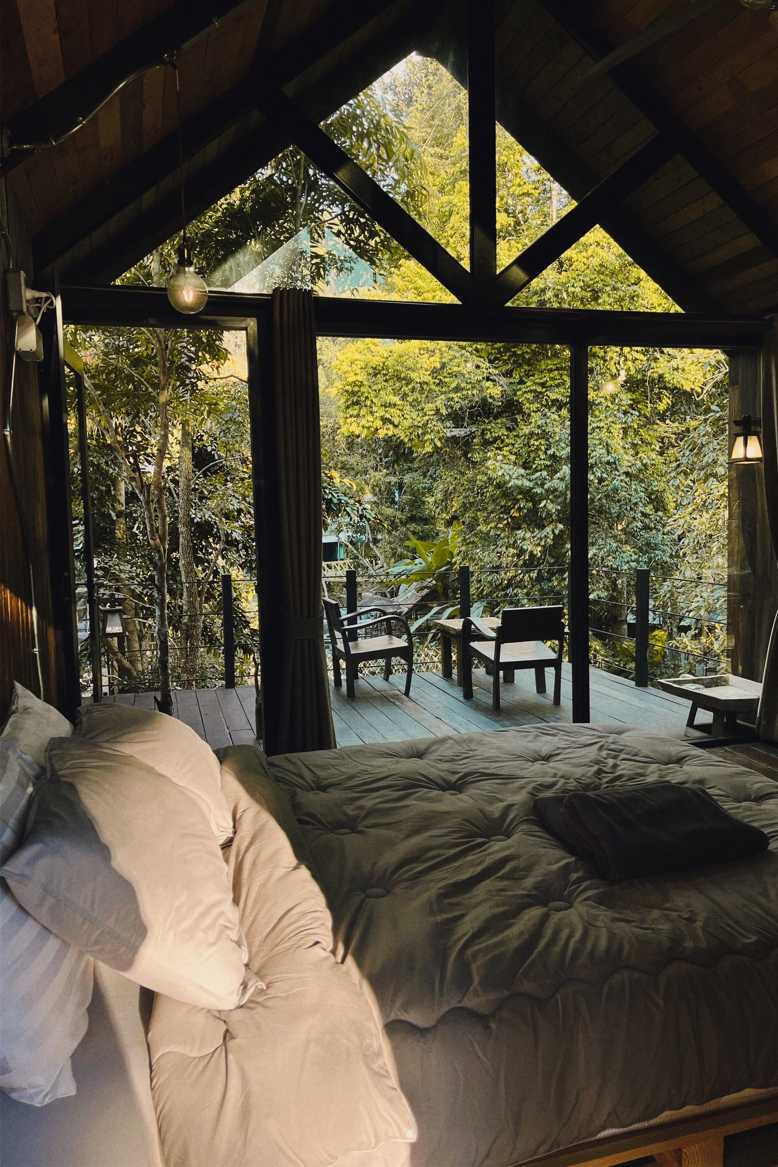 Inside a cozy bedroom with large windows overlooking a lush green forest and a small balcony with chairs.