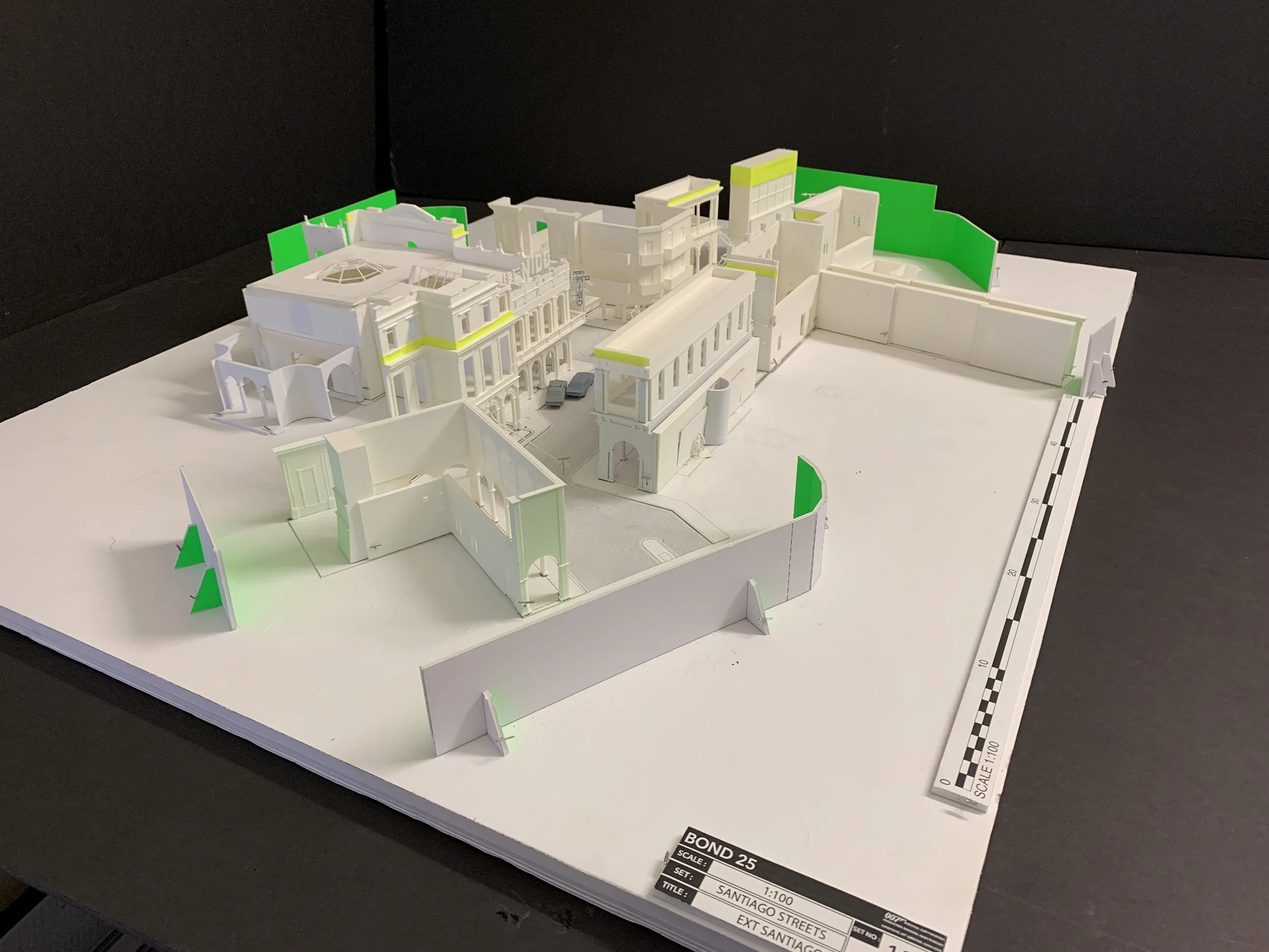 Architectural model of a building complex with multiple structures, parking spaces, and surrounding walls, displayed on a white base with a scale ruler along the side.
