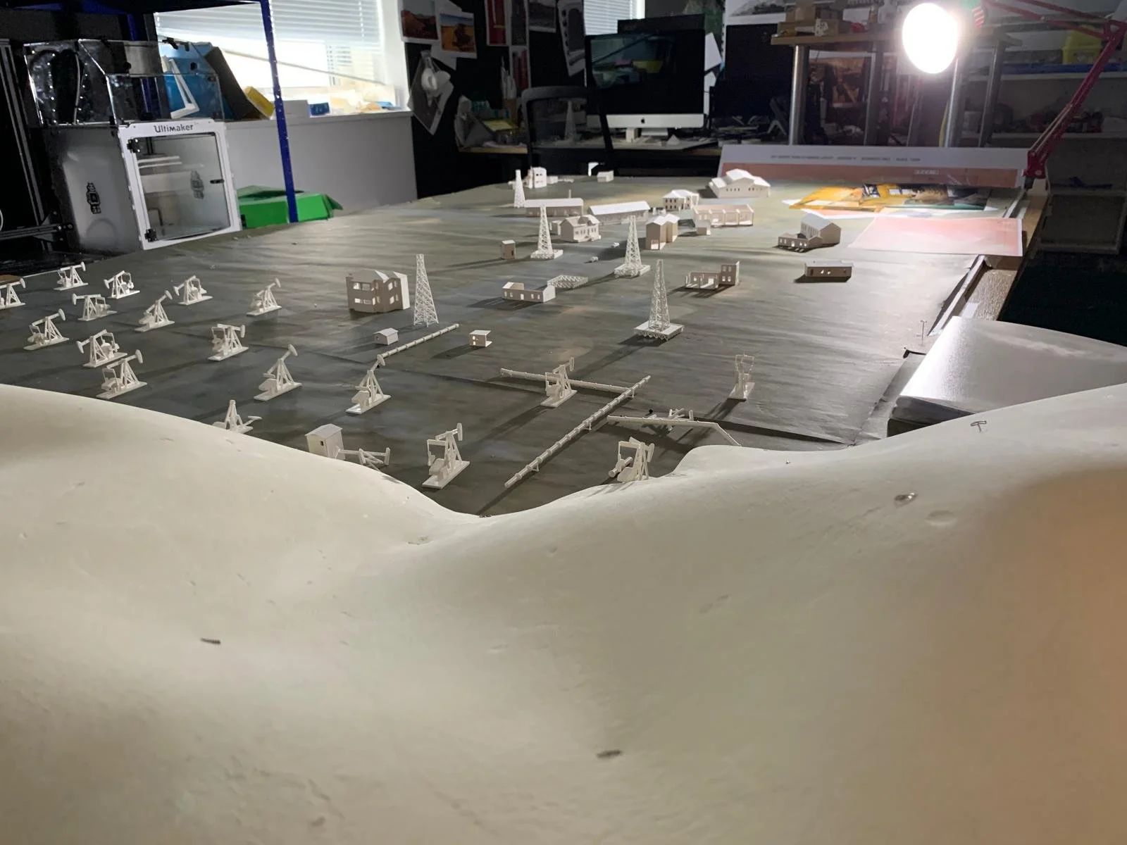 A model of an oil field with oil rigs, buildings, and electrical towers on a table in a workshop or studio.