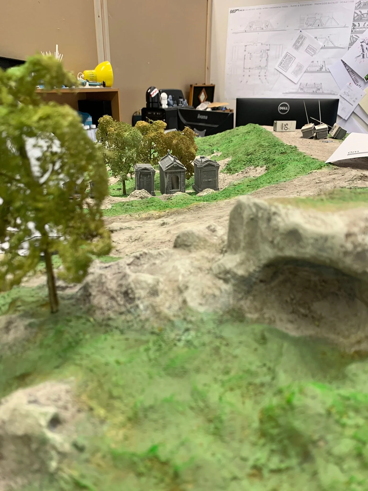 Miniature landscape with trees, rocks, and small structures resembling tombs or monuments, set on a terrain with green and beige textures, with an office workspace in the background.