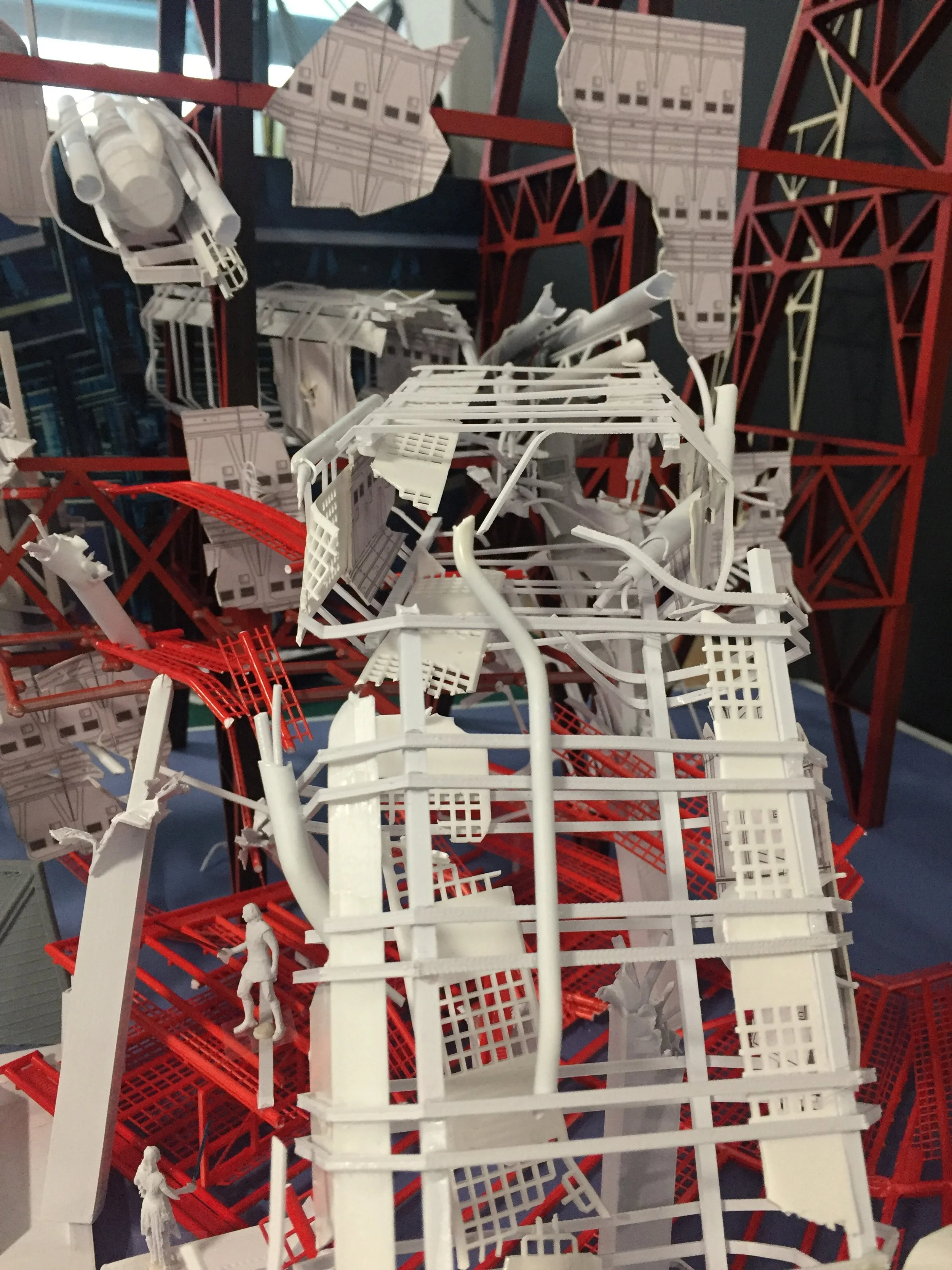 Miniature architectural model of a building with white and red structural elements, small figurines, and architectural plans.