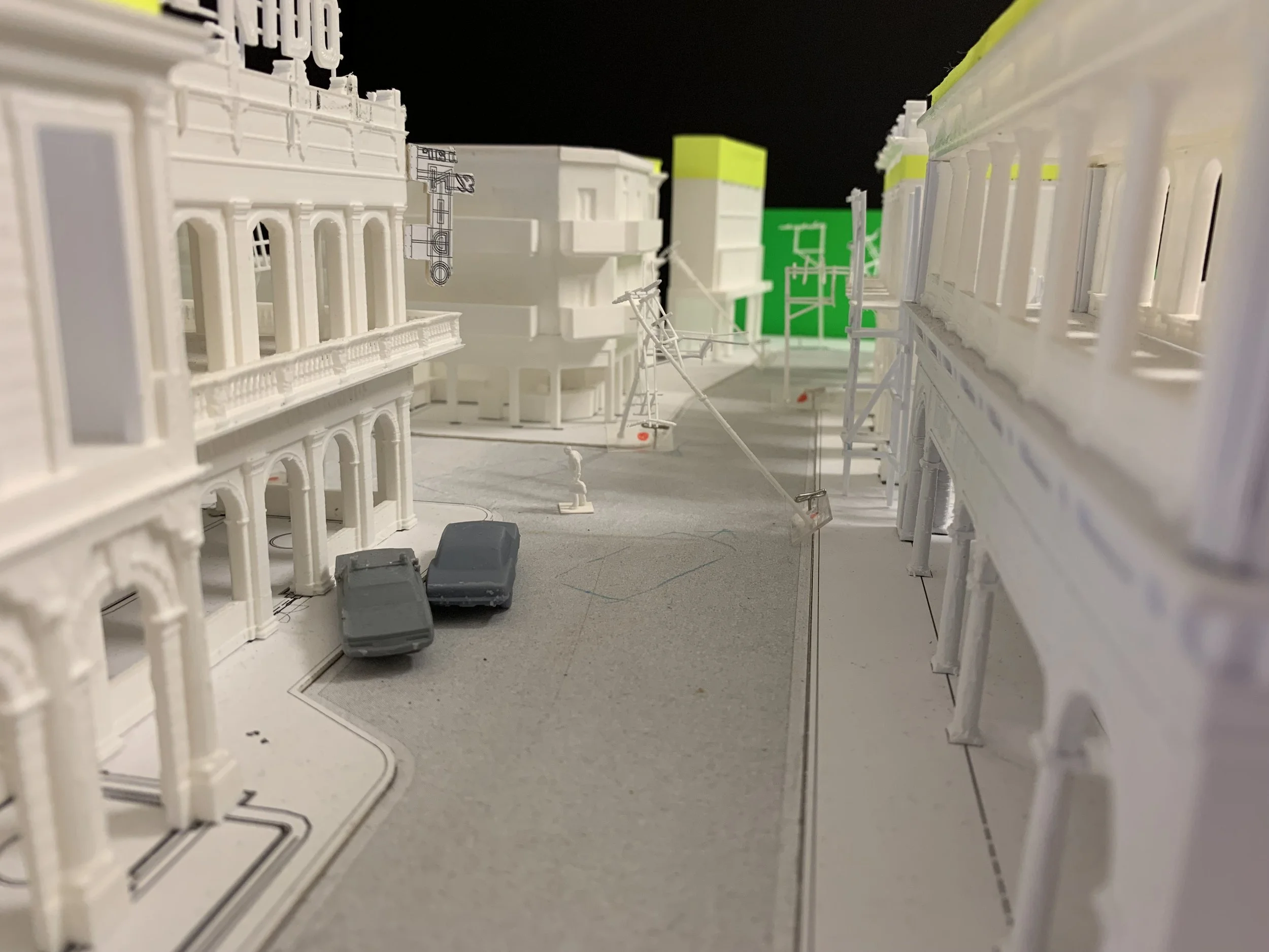 Scale model of a city street with white buildings, parked cars, and construction scaffolding, with a green backdrop in the background.