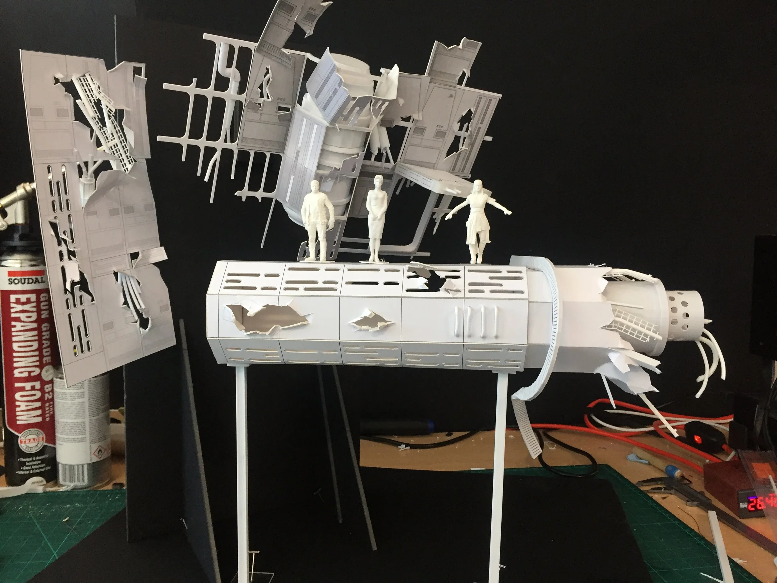 A detailed white model of a spaceship or space station, with miniature figures of people on it, set against a black background with tools and a fire extinguisher nearby.