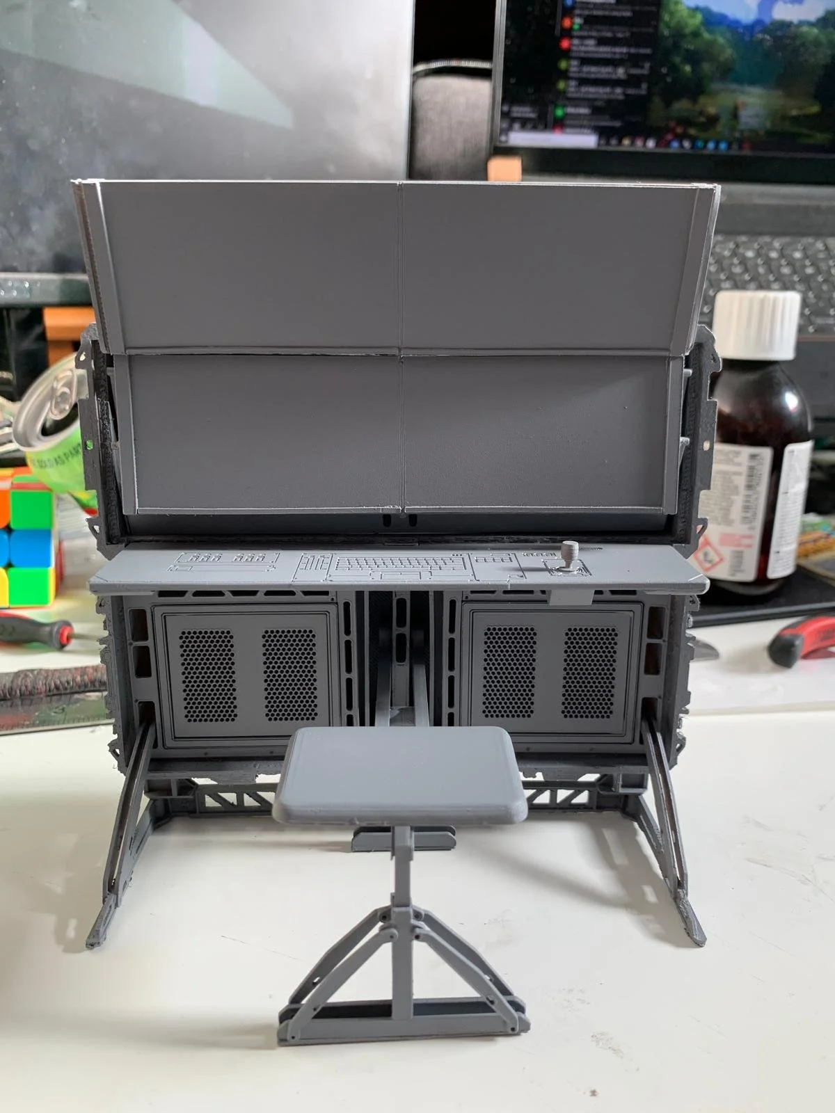 Gray open miniature model of a computer setup with a chair, set on a white surface, with assorted items and a laptop in the background.