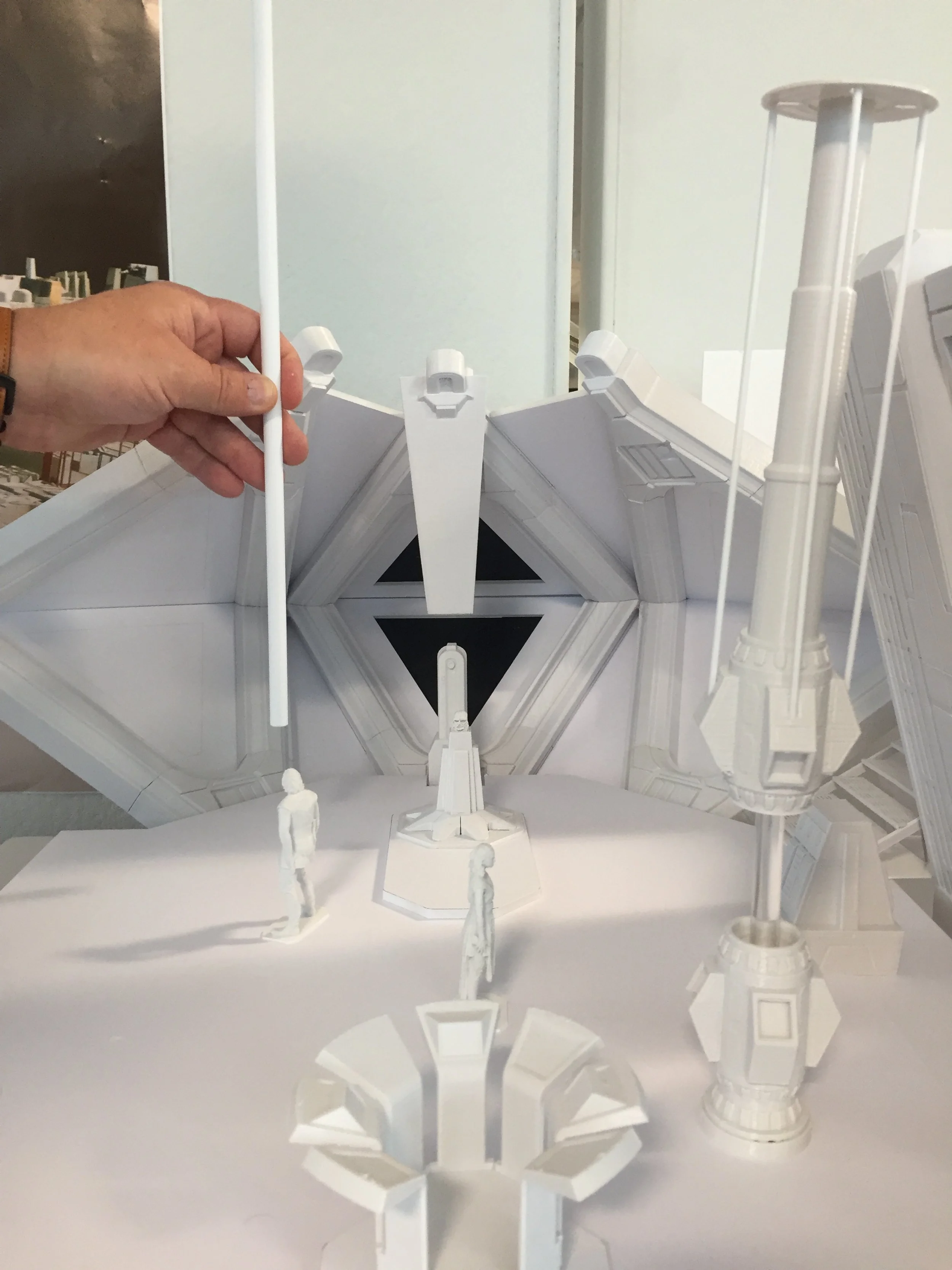 A person holds a white rod in front of a detailed white model of a sci-fi or spaceship scene, which includes miniature human figures, a large structure, and various mechanical or technological elements.