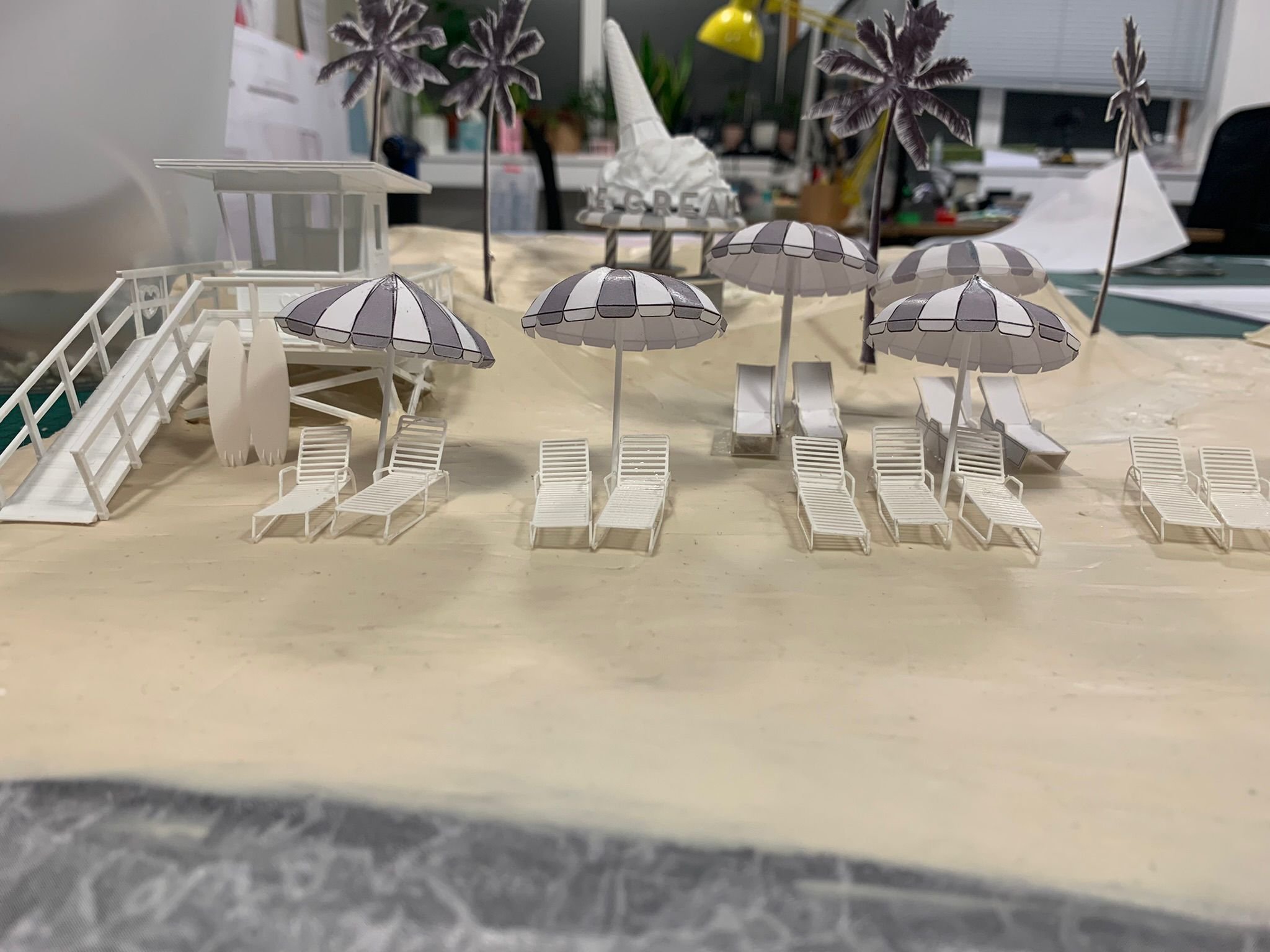A miniature model of a beach scene with white lounge chairs, umbrellas, and palm trees on a sandy surface with an office in the background.