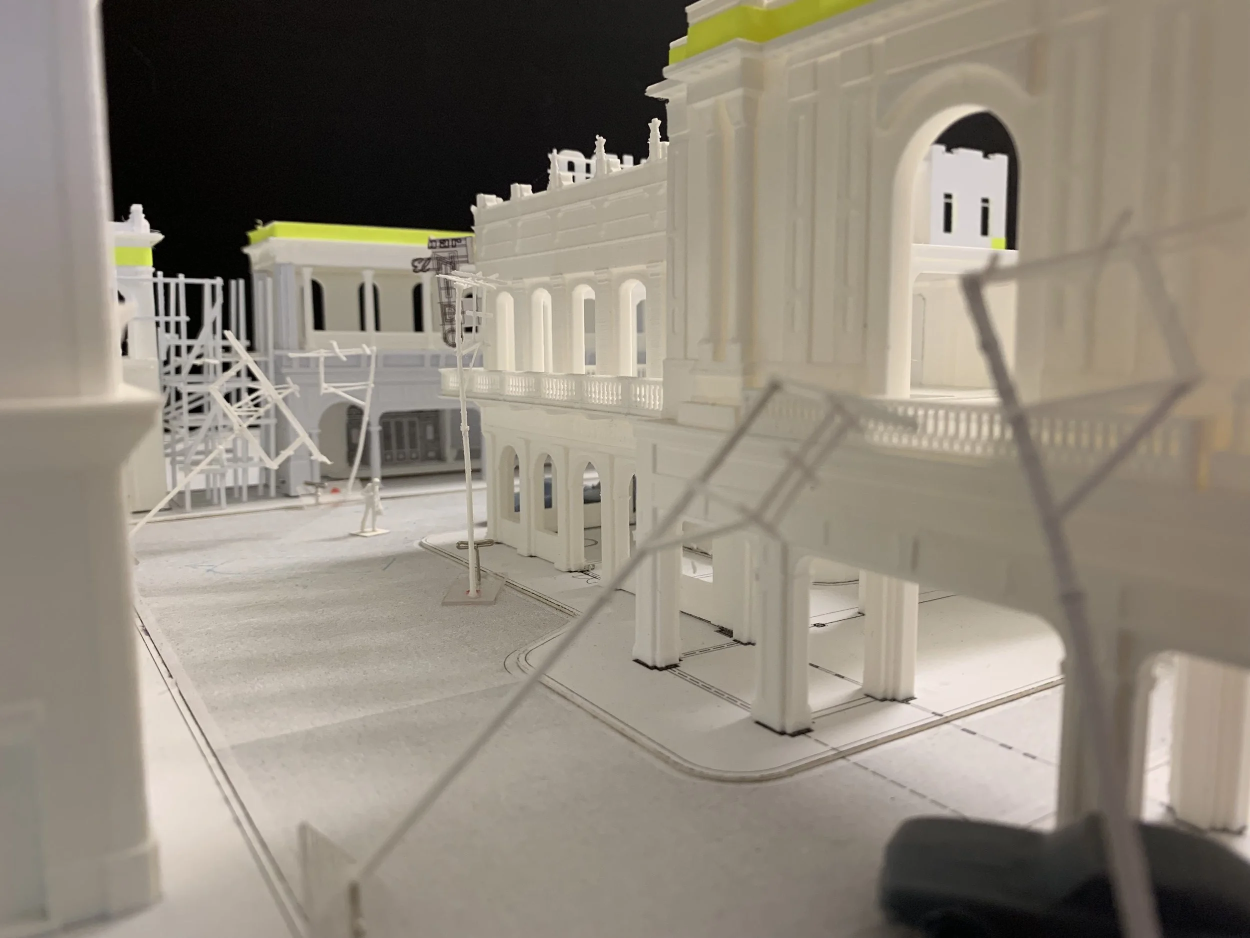 Architectural model of a white building with multiple arches and balconies, surrounded by small street lamps and fencing.