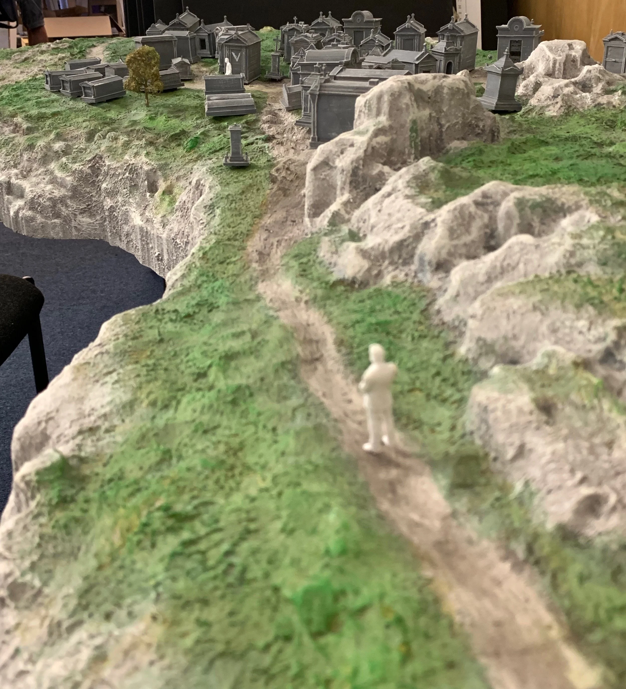 A miniature landscape model with a winding dirt path, rocky terrain, and green grass. There is a tiny figurine of a person standing on the path and small tombstones or mausoleums in the background.