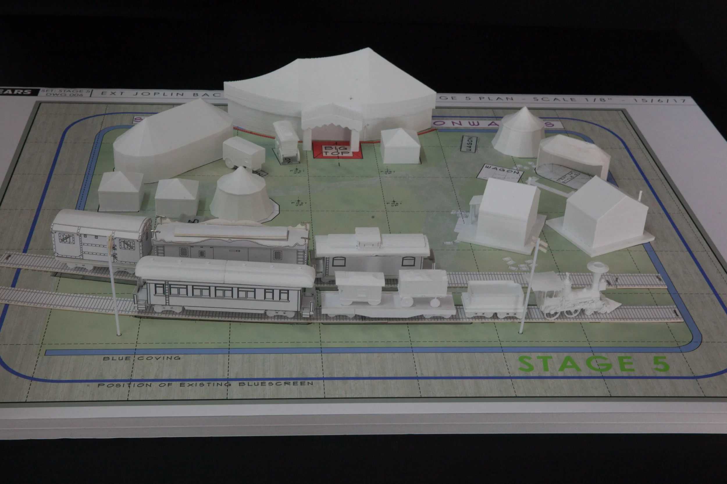 Architectural model of a train station with miniature buildings and a train on a track, placed on a layout plan labeled 'Stage 5'.