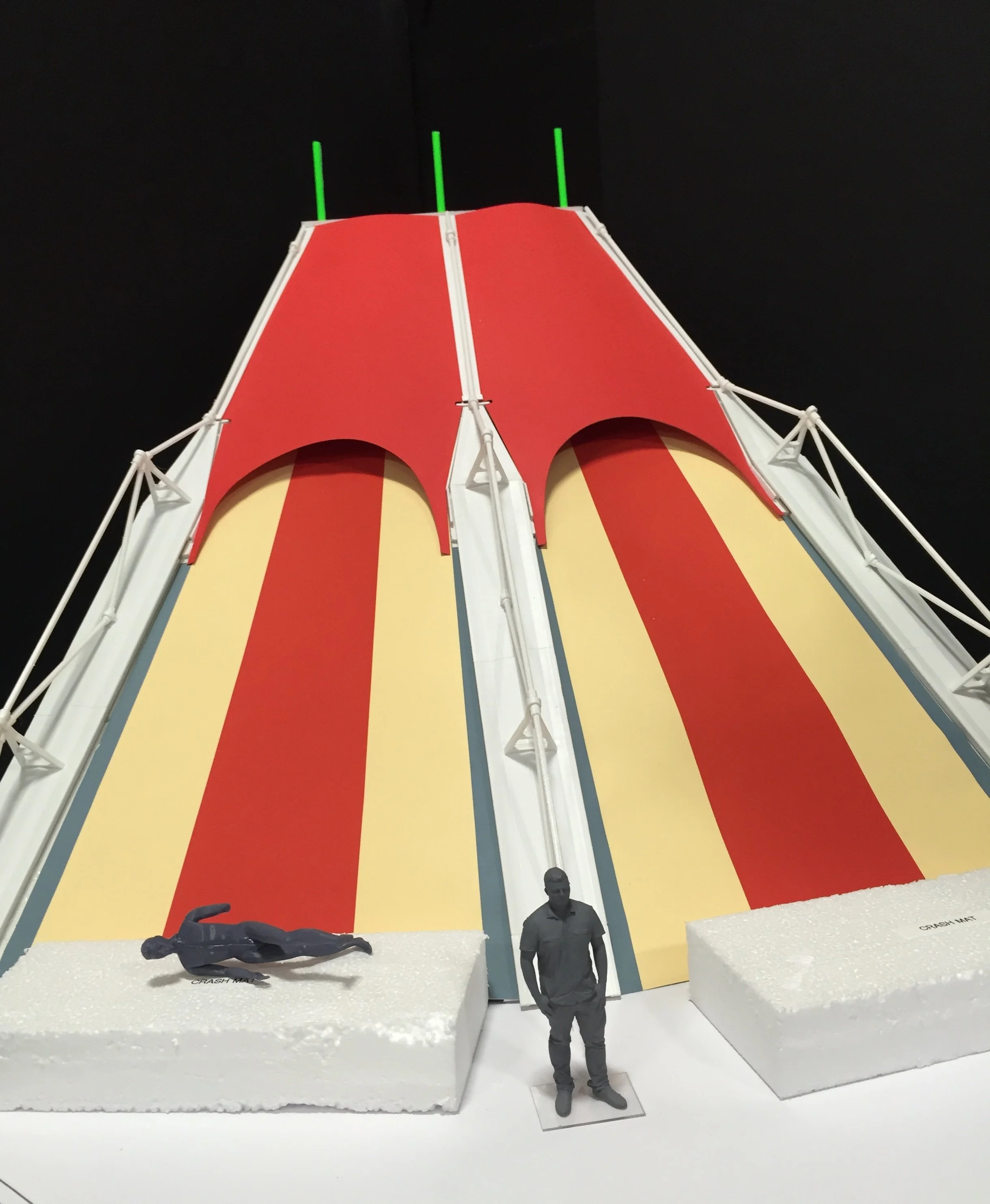A scale model of a carnival ride with a large red and yellow striped slide, two foam crash dummy figures, and a miniature person standing at the base, against a black background.