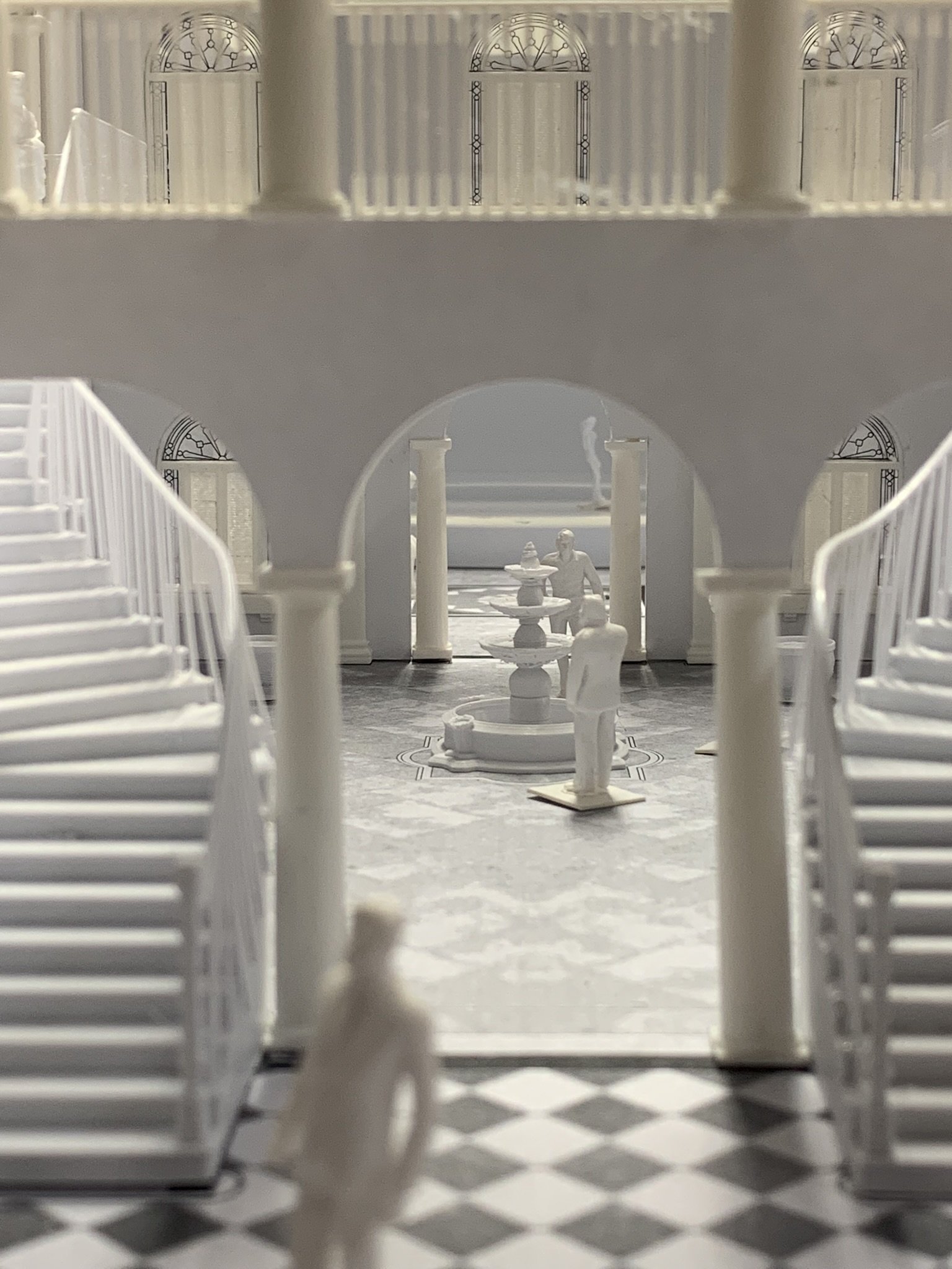 Miniature white architectural model of a two-story building with stairs, columns, sculptures, and a fountain in a black and white checkered floor setting.
