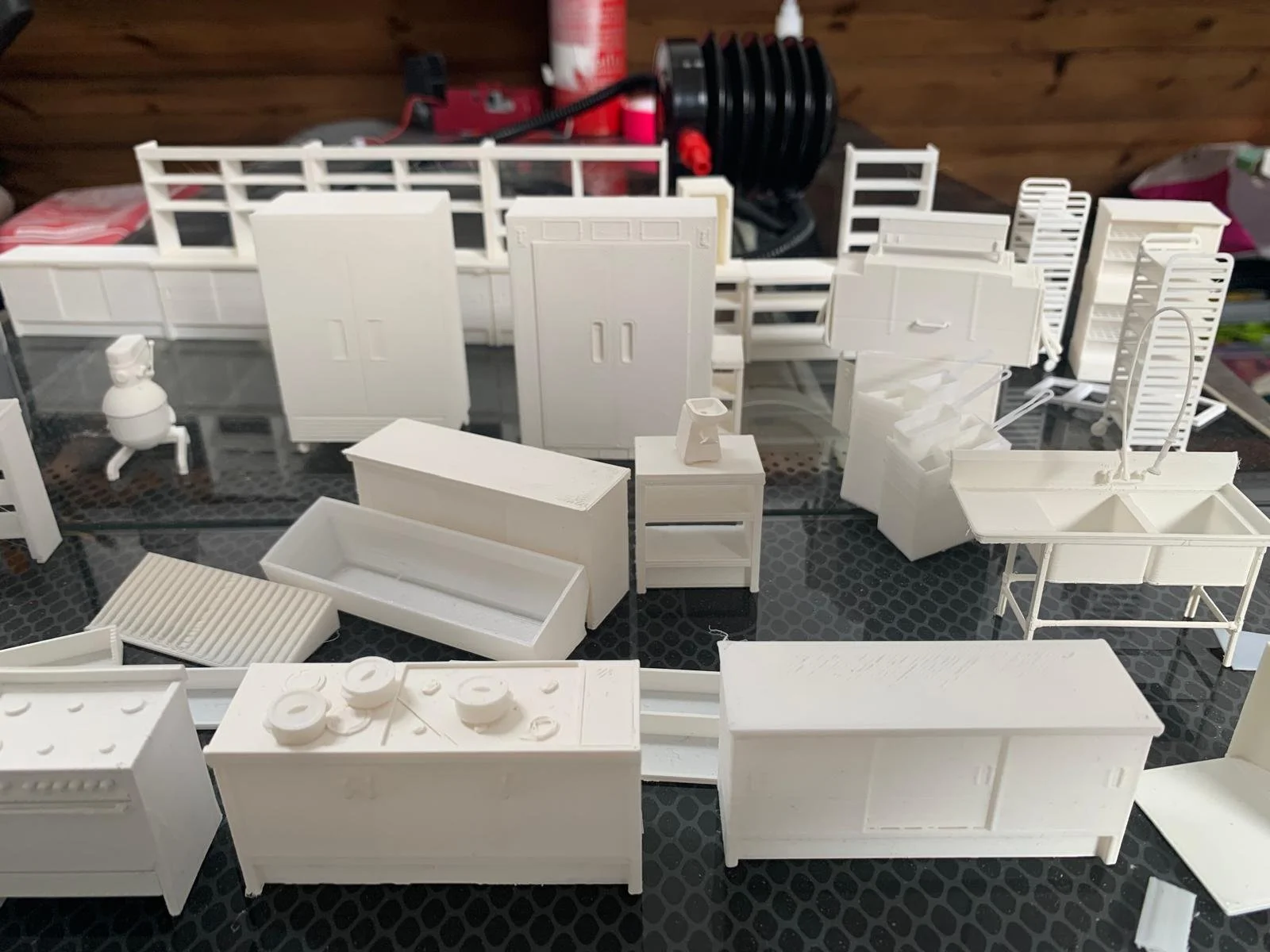 Photo of miniature white model furniture pieces arranged on a black surface, including cabinets, tables, and appliances, with a wooden wall and some objects in the background.