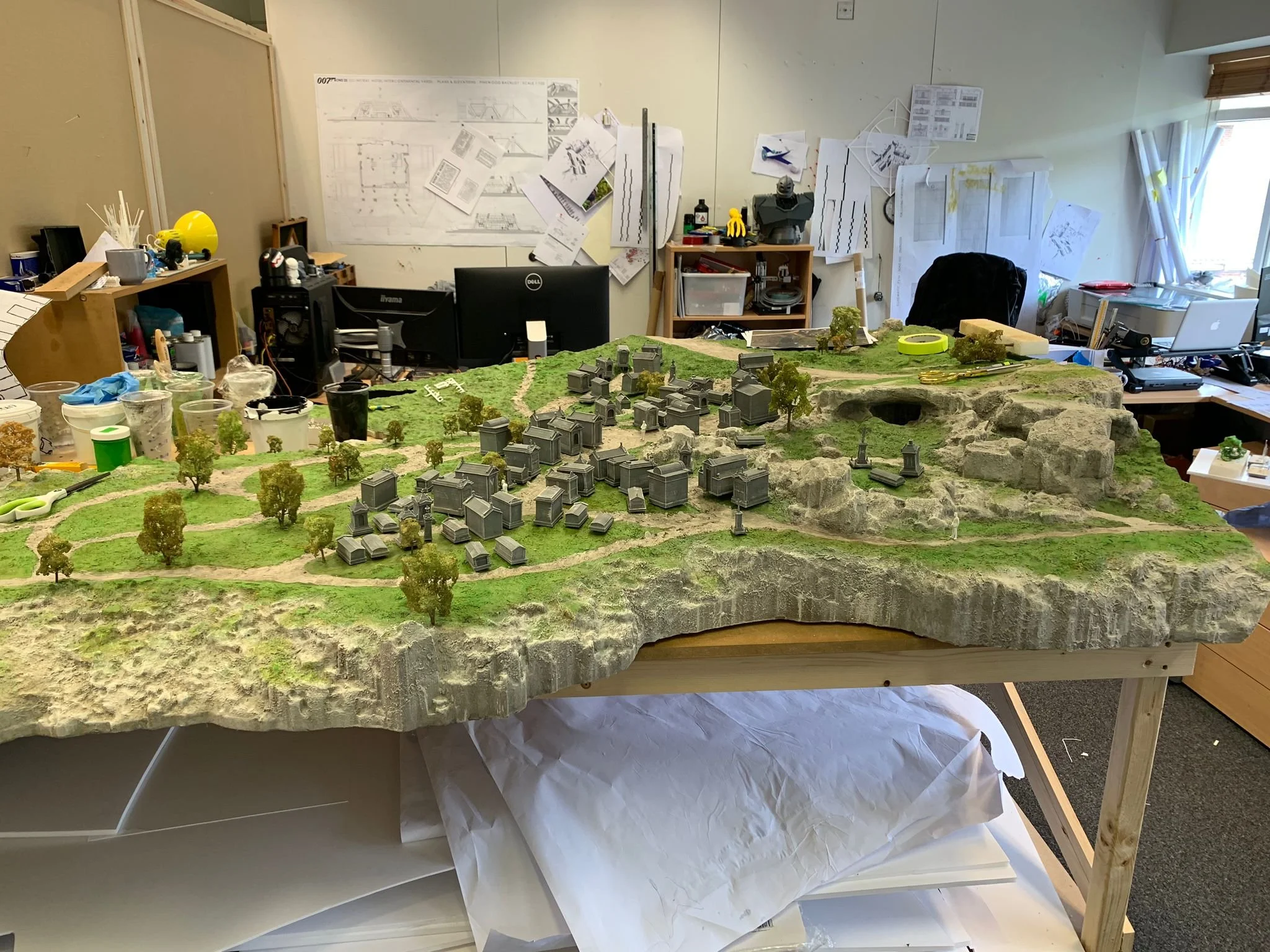 A detailed miniature landscape model with small trees, buildings, and rocky terrain on a table in a workshop or office setting.