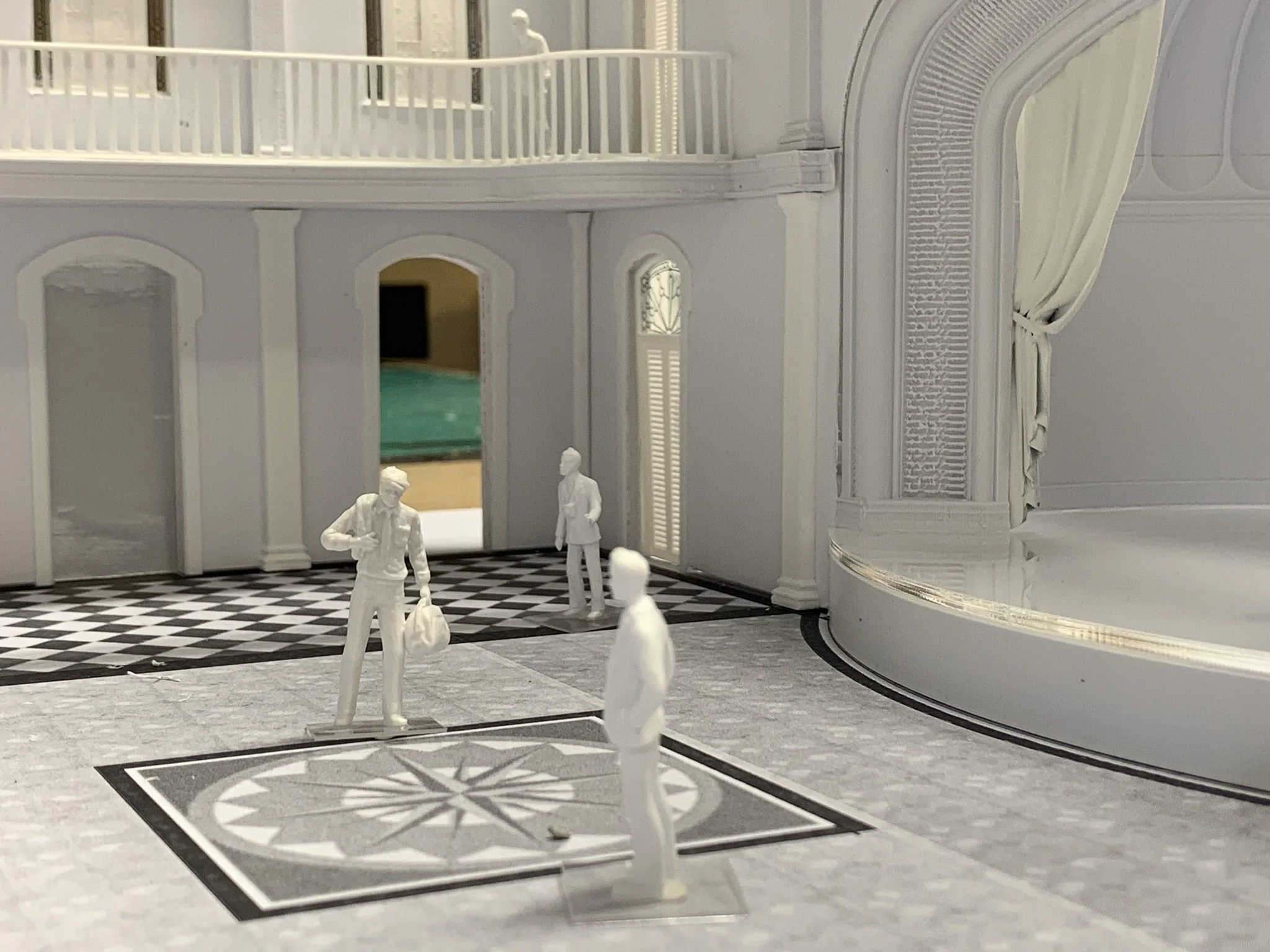 Miniature model of an elegant interior with three white figurines representing people, a checkered floor, and ornate architectural decor.