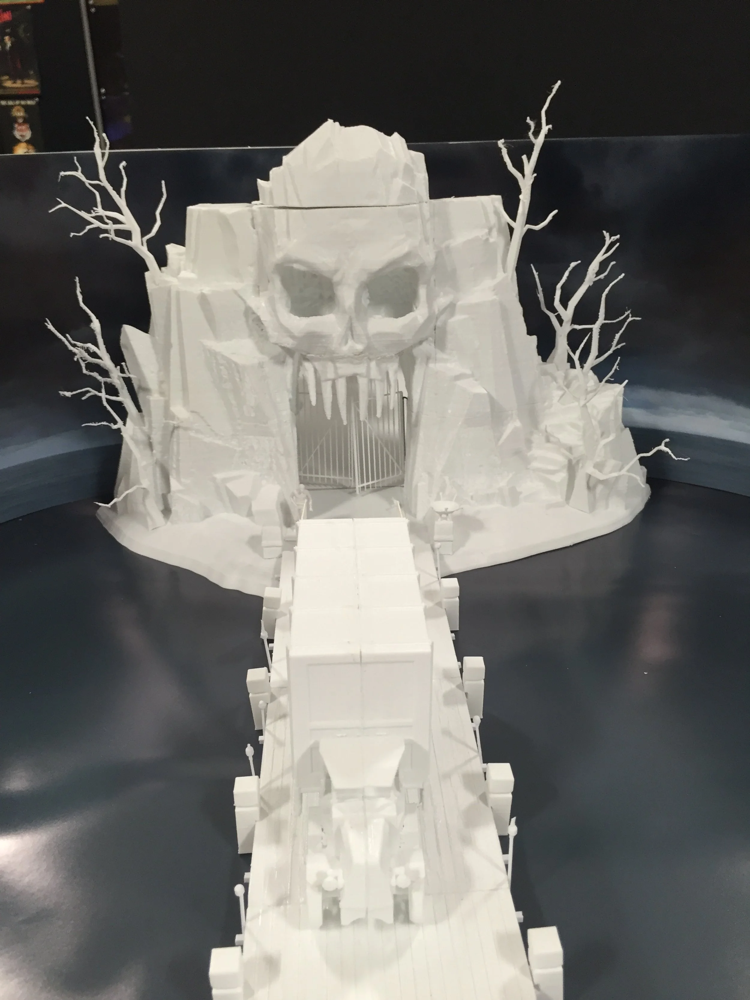 White paper model of an icy mountain with a large skull face carved into the cliffs, featuring dark eye sockets and sharp teeth, with leafless twisted trees on either side, and a bridge leading to the skull's gaping mouth.