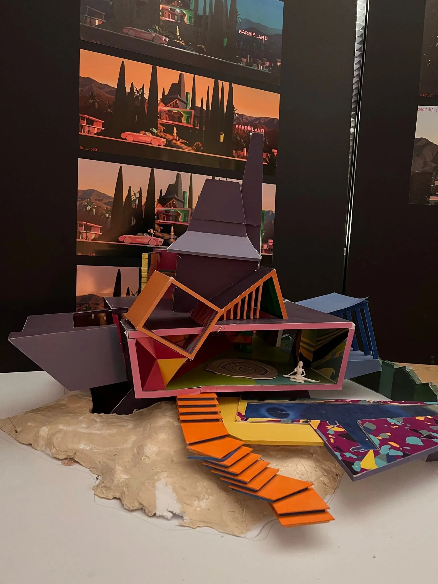 Colorful architectural model featuring multiple levels, ramps, and geometric shapes, placed on a textured base with a black background and posters depicting a sunset scene with trees and mountains.