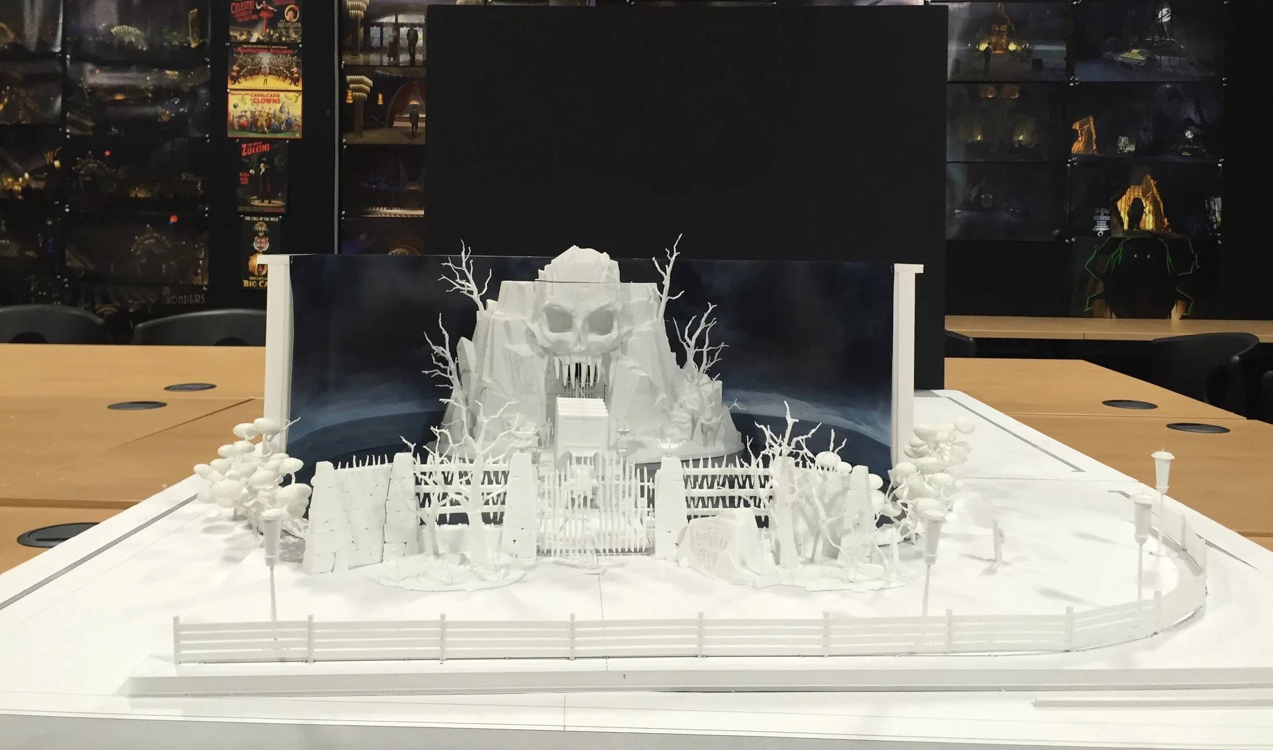 White architectural model of a haunted house with a skull-shaped facade, surrounded by trees and a fence, set on a table in a room with posters in the background.