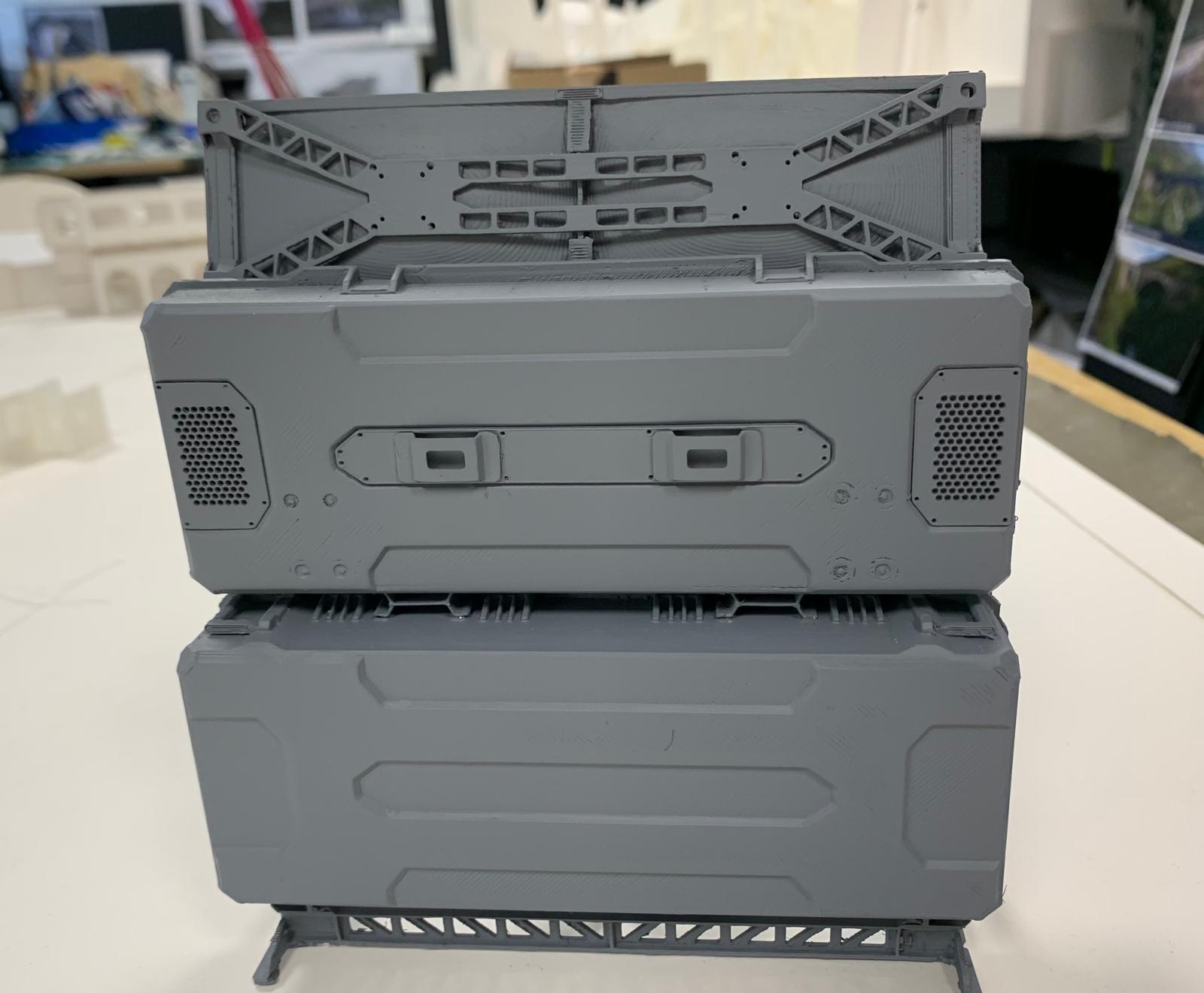 Stacked gray plastic model spaceship or robot parts on a white surface, with a workshop or storage area in the background.