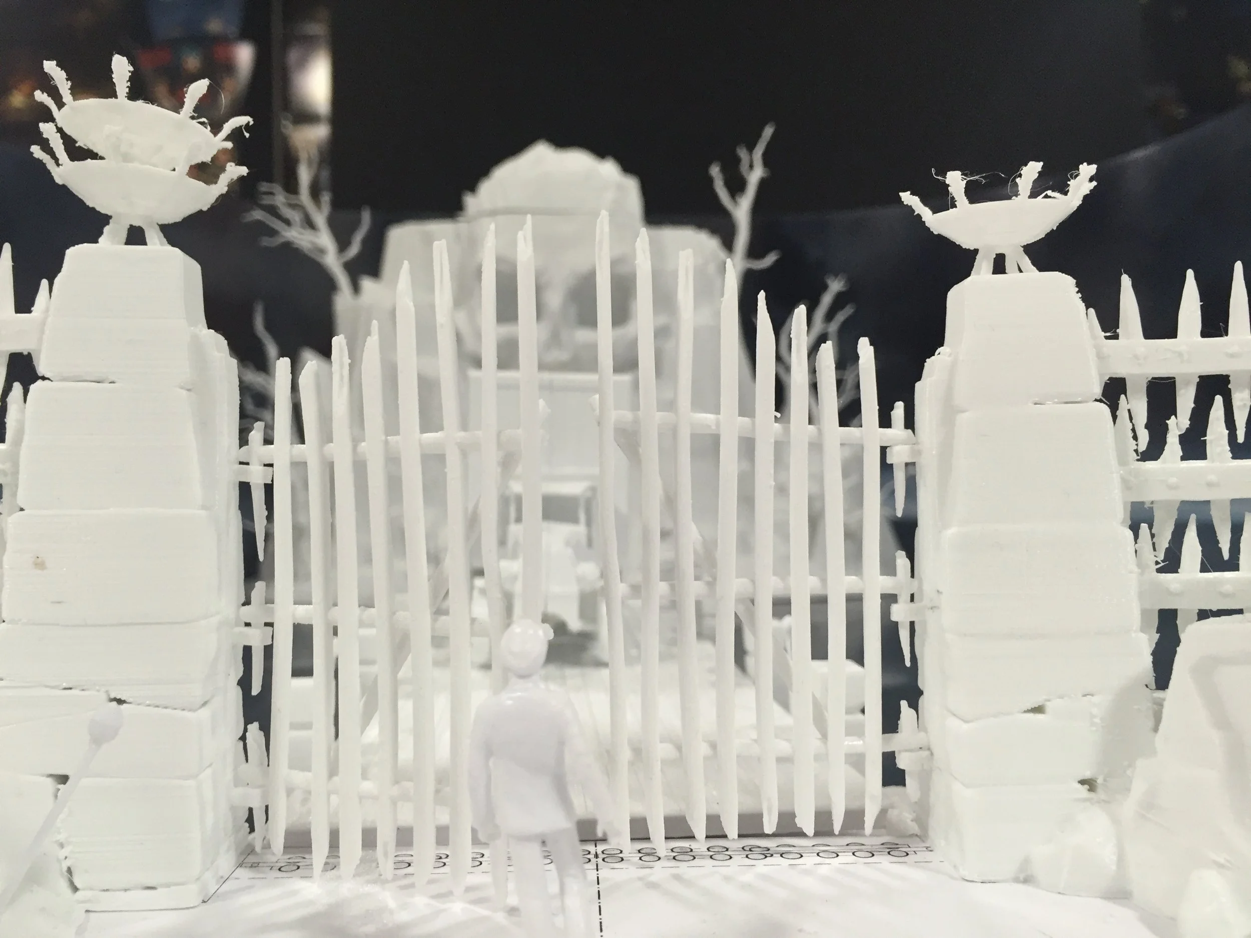 A white miniature model of a gated entrance with two tall pillars topped with decorative sculptures, and a small figure standing in front of the gate.