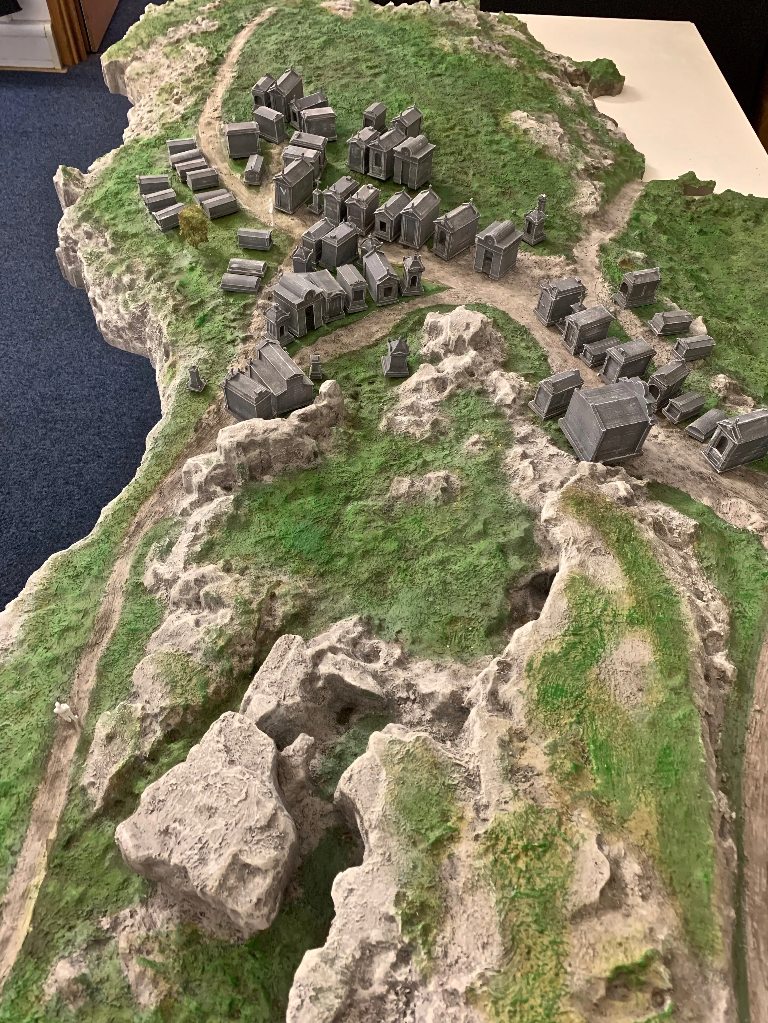 A detailed miniature landscape model depicting a small town with buildings, roads, and surrounding terrain with grassy and rocky areas.