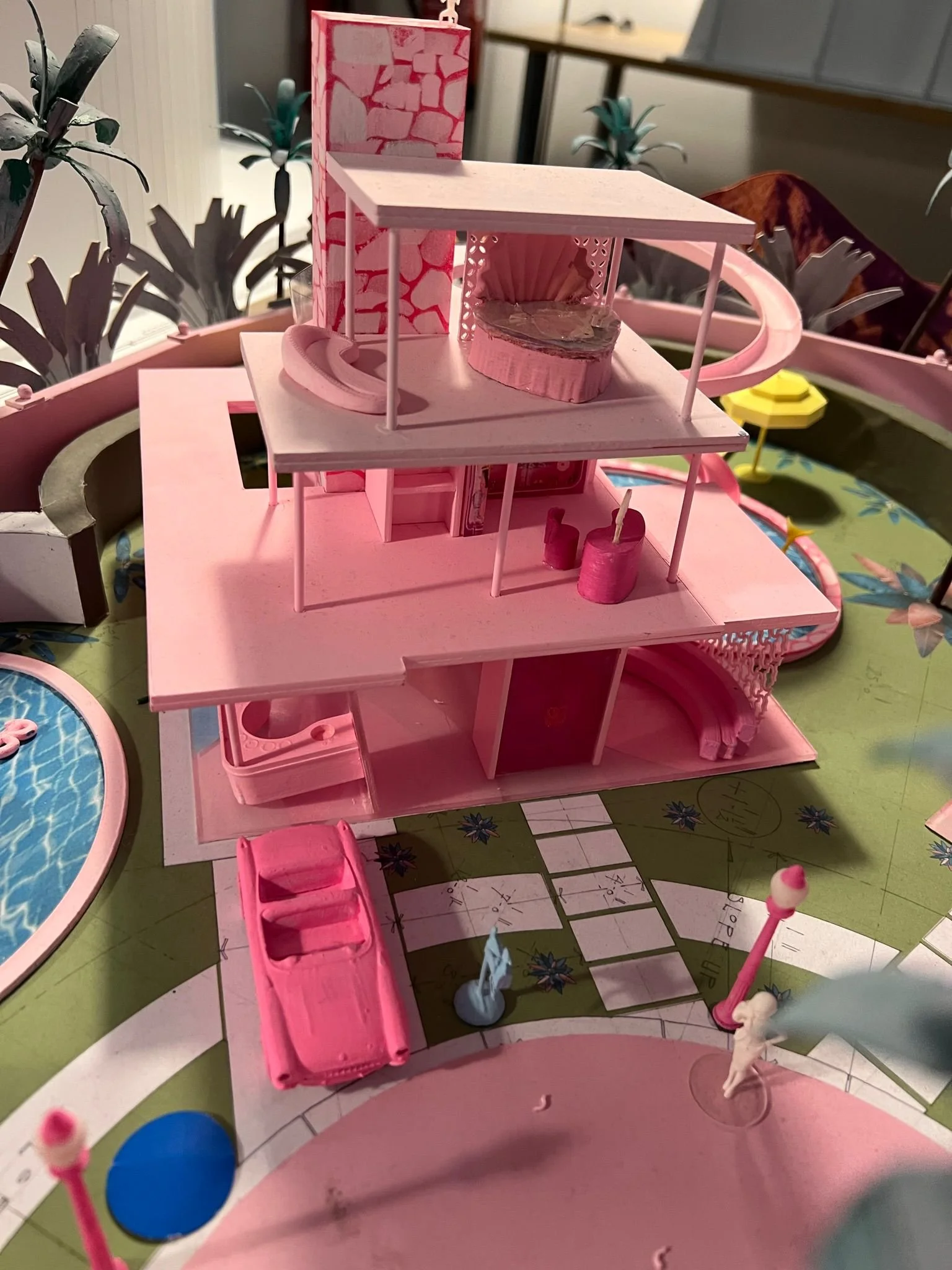A pink plastic dollhouse with multiple floors, a car parked in the driveway, and various miniature accessories inside and around it, set on a colorful game board.