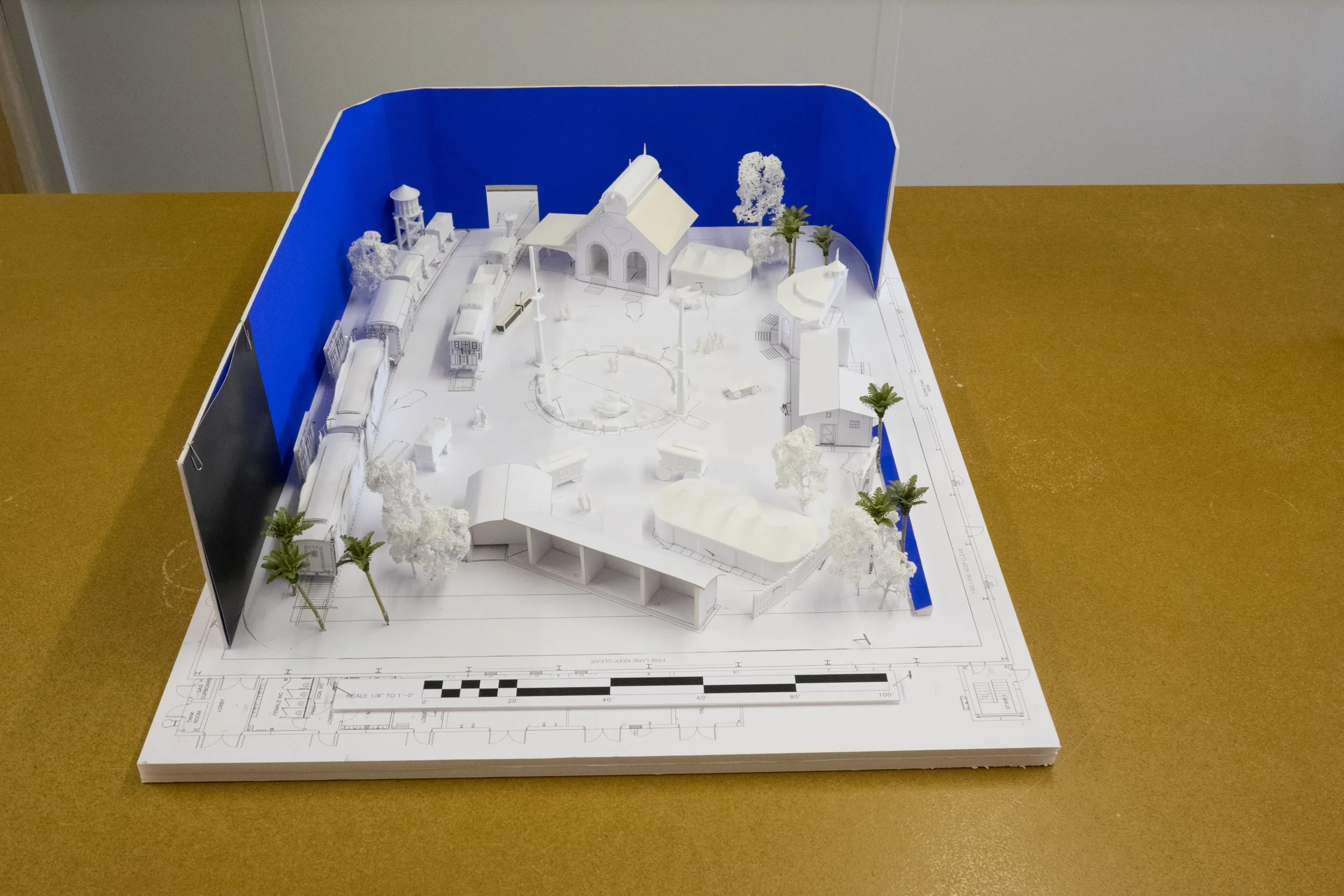 A scale model of a park or village with small buildings, trees, and a train, set on an architectural blueprint base, with a blue and black backdrop on one side.