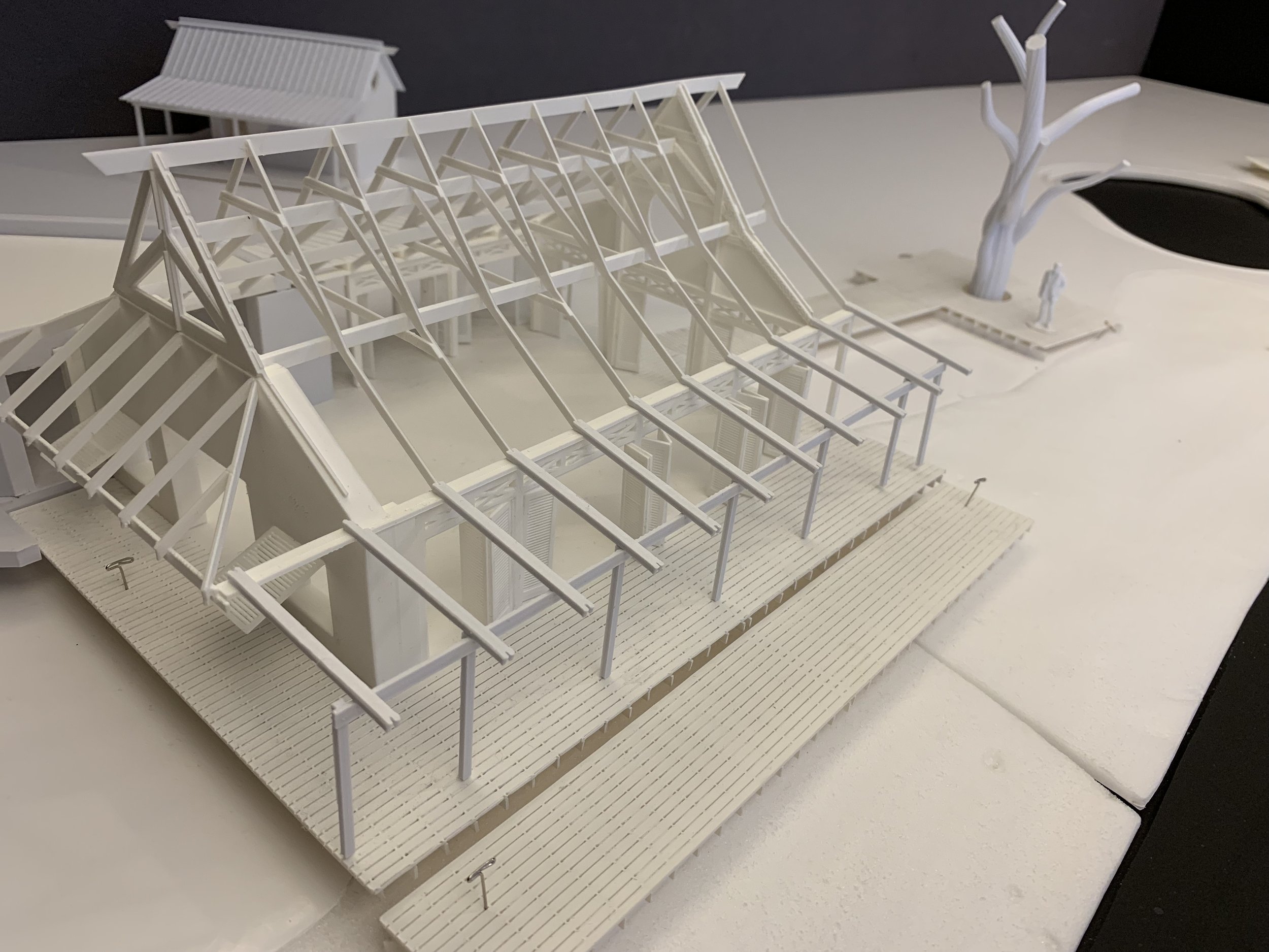 Architectural scale model of a building with a sloped roof and an outdoor deck, featuring a leafless tree and small figures, on a white base.