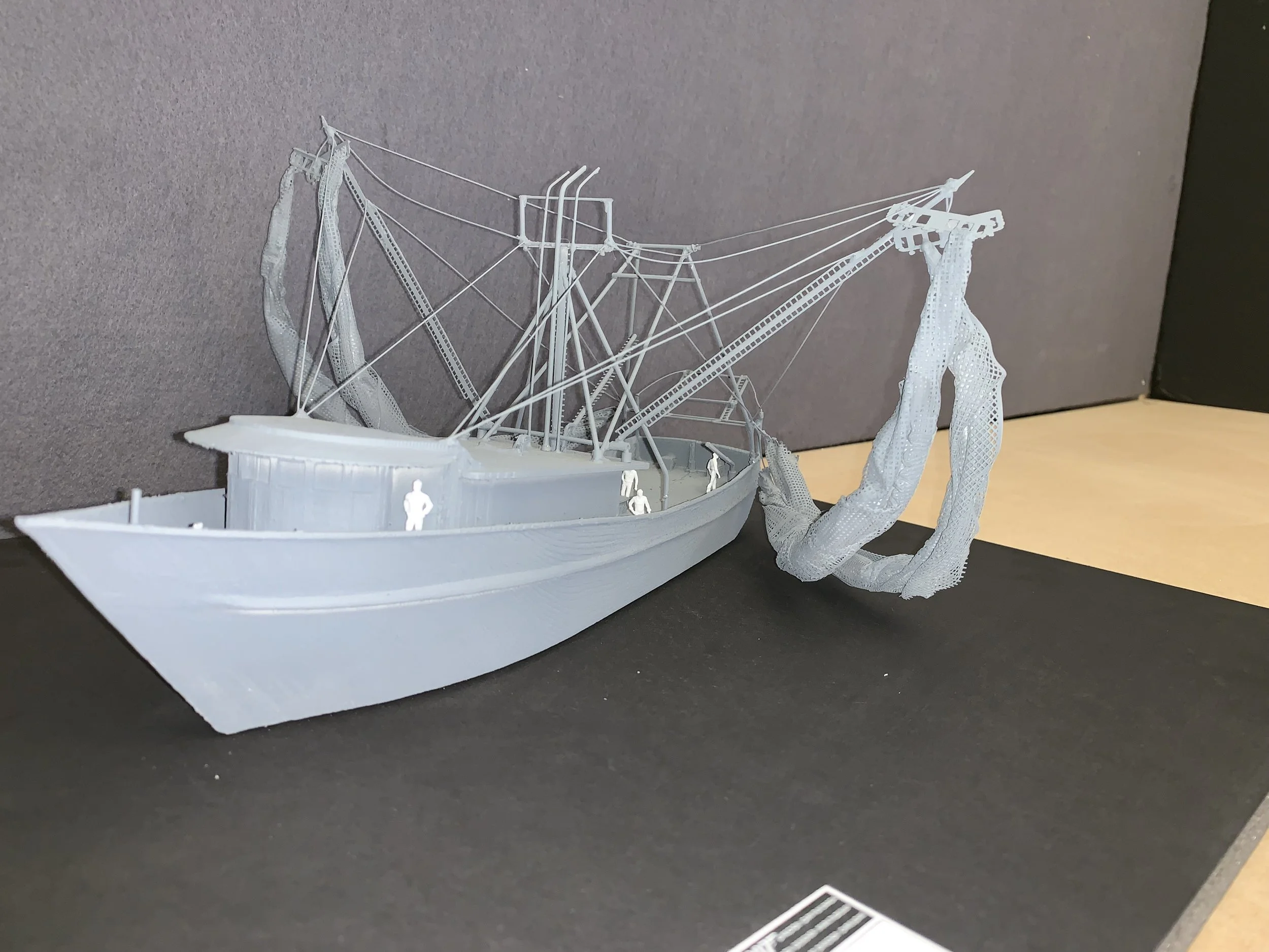 A detailed scale model of a boat with a crew of miniature figures, featuring rigging and a large net or fabric hanging from the mast, displayed on a black surface against a gray backdrop.