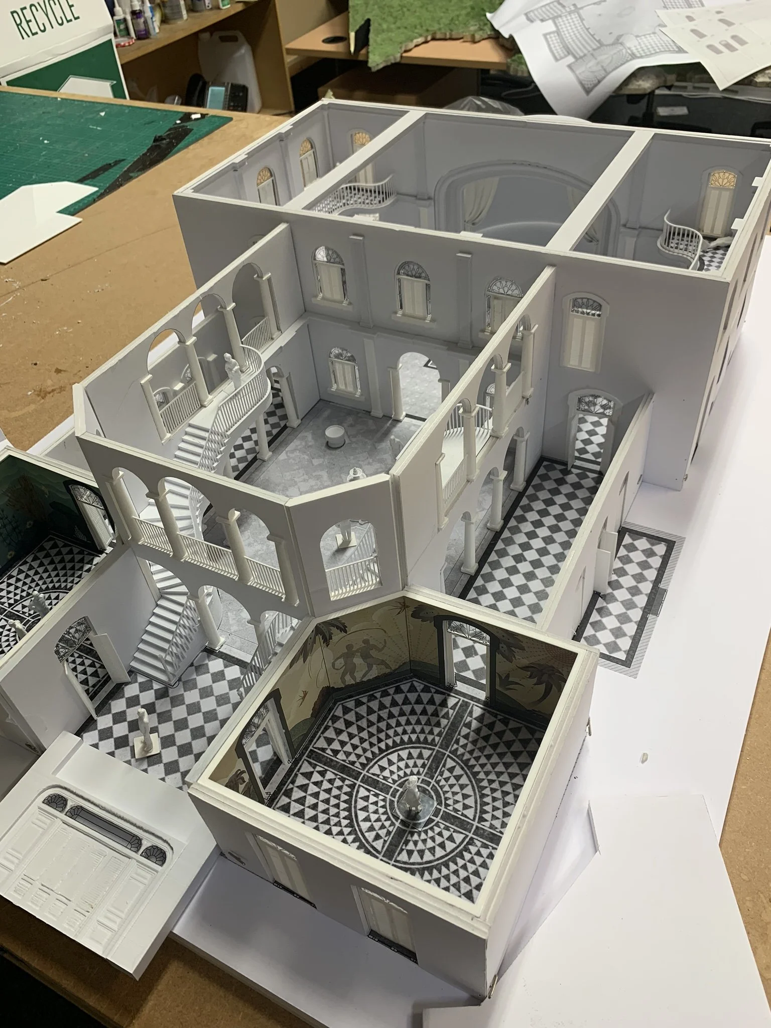 A detailed architectural model of a mansion with multiple rooms, balconies, and a patterned black and white tile floor, assembled on a worktable.