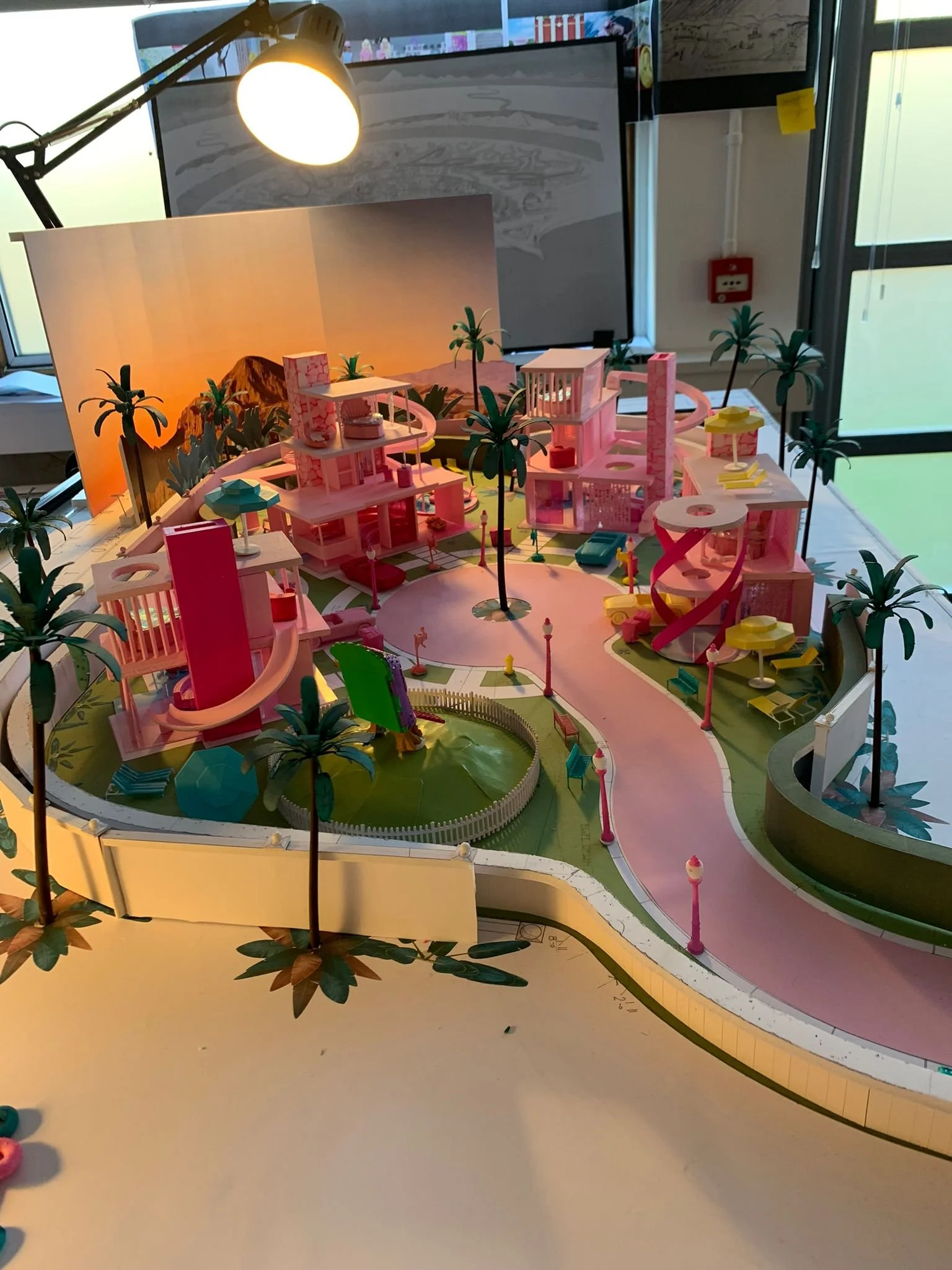 A colorful architectural model of a tropical urban community with pink buildings, green trees, and winding roads, displayed indoors under a bright light.