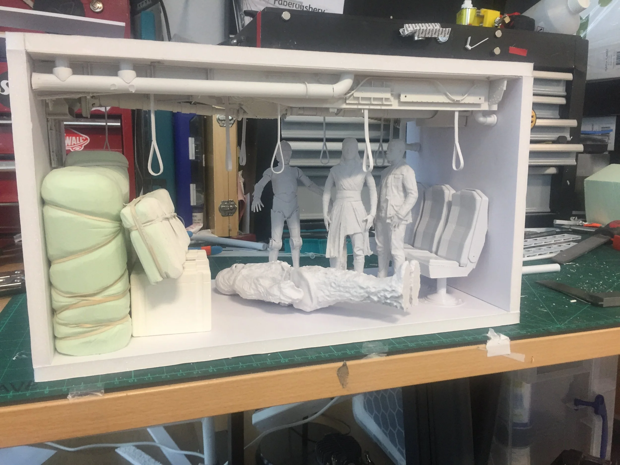 Miniature sculpture of three figures sitting in a bus, with one figure standing. The sculpture is in a diorama with seating and hanging straps, on a workbench surrounded by tools and supplies.