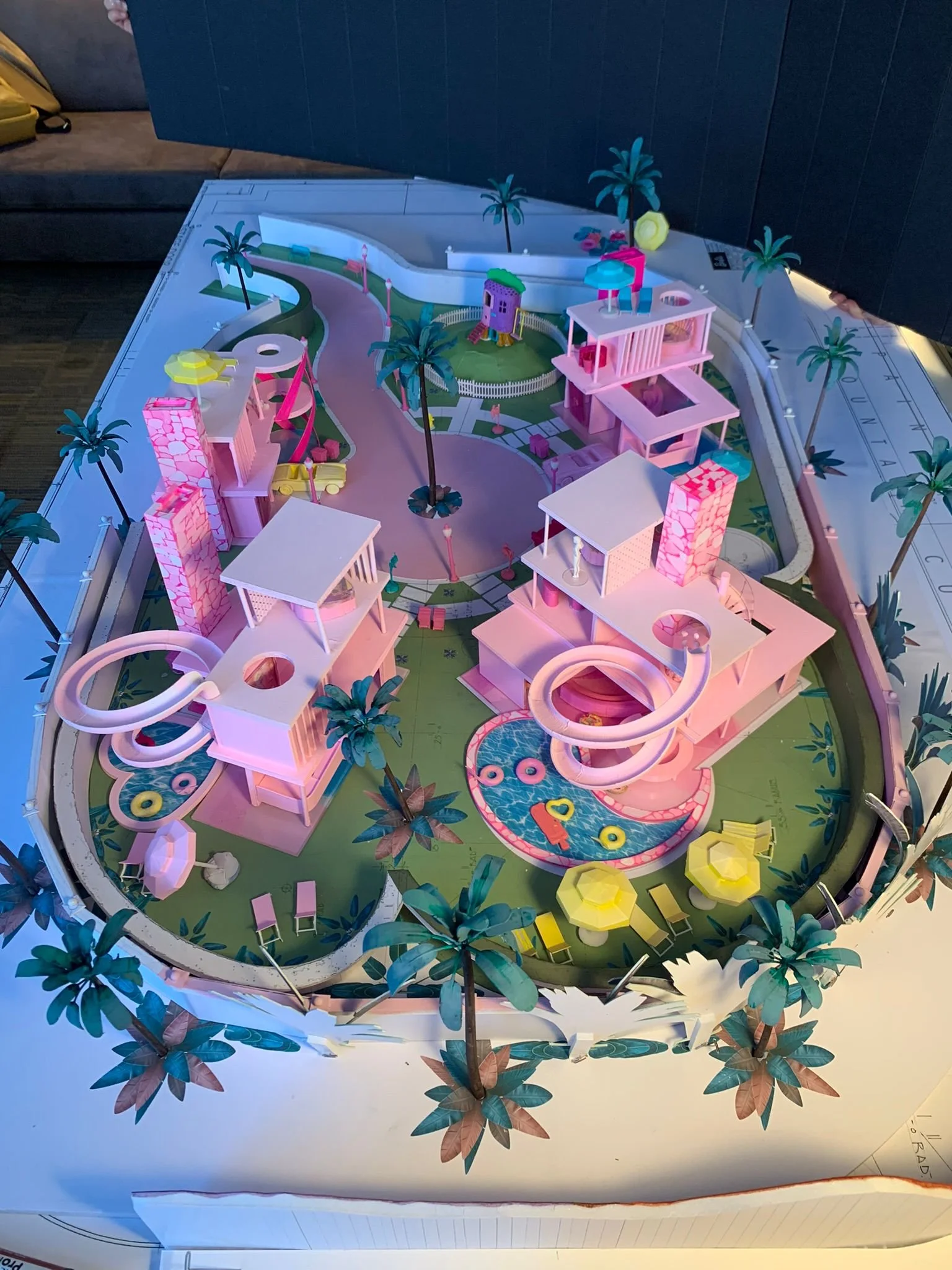 A colorful architectural model of a modern tropical resort with pink buildings, palm trees, water features, lounge chairs, and winding slides on a large outdoor canvas.