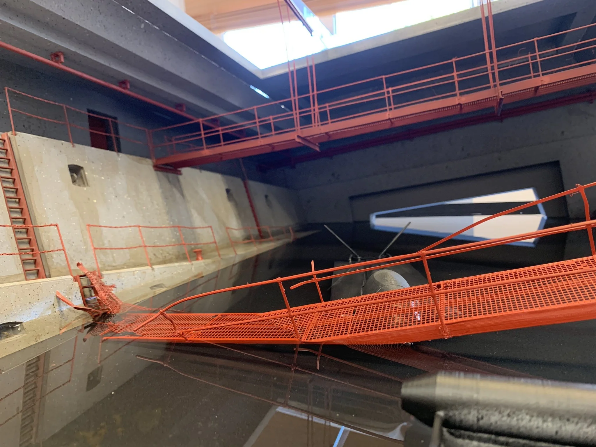 A model of an industrial or architectural structure featuring an orange metal walkway with railings and a ladder, surrounded by concrete walls with small openings, and a black rectangular opening with a white geometric shape inside.