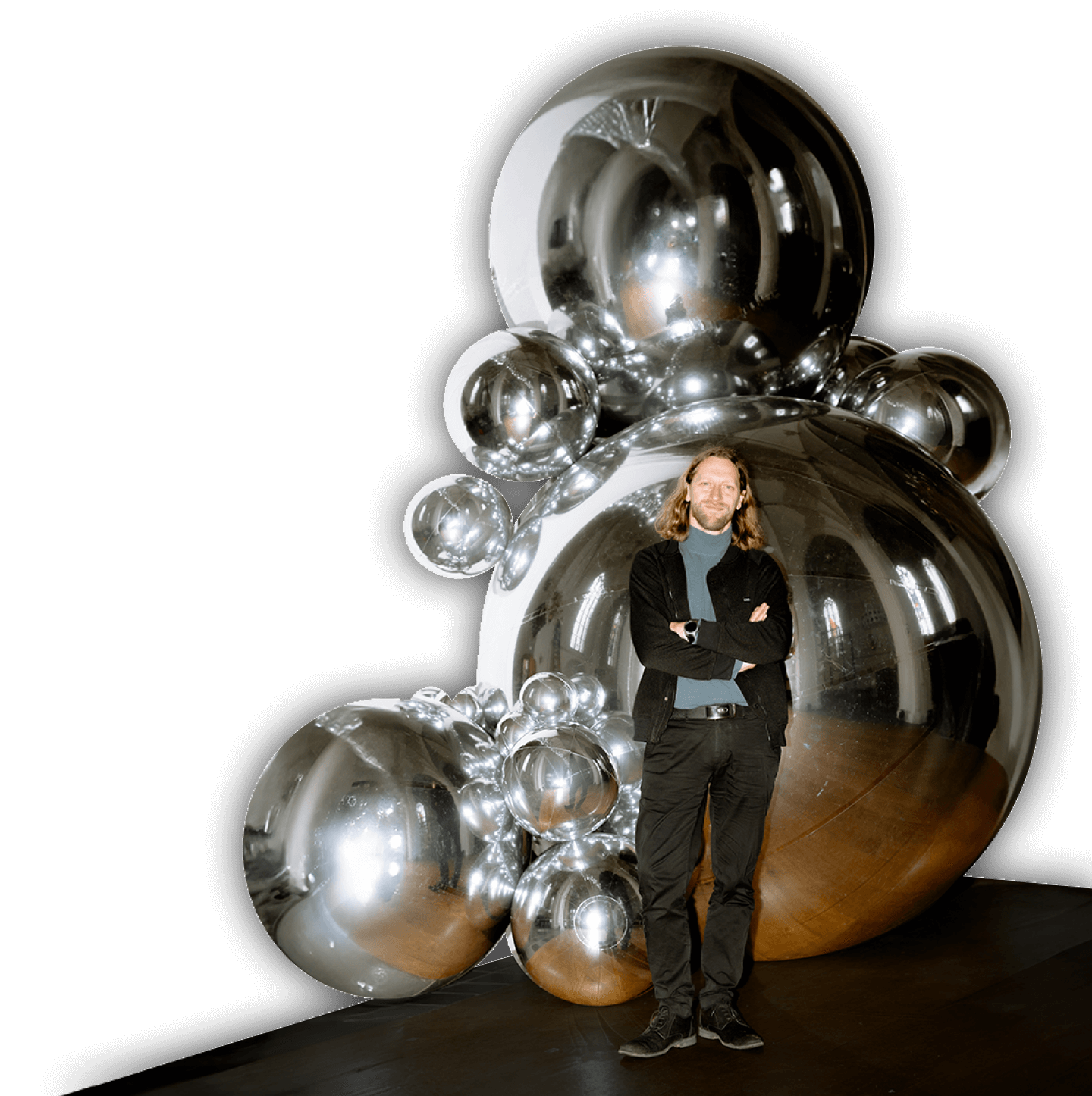 Ein Mann steht vor einer Skulptur aus großen, glänzenden, silberfarbenen Kugeln. Der Raum ist dunkel, und die Kugeln reflektieren die Umgebung.
