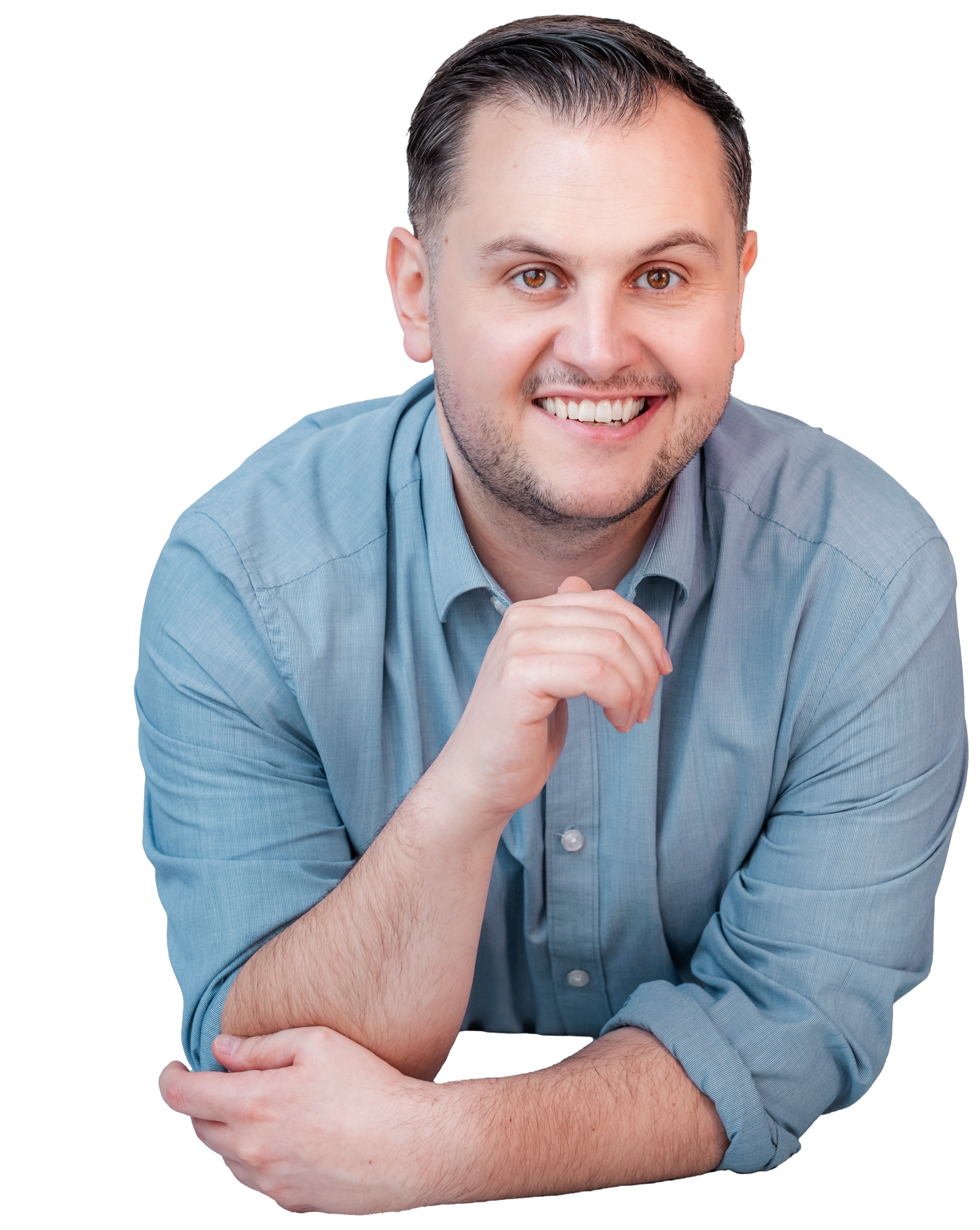 A smiling man in a light blue button-up shirt, resting his chin on his right hand with his left arm folded on a surface.