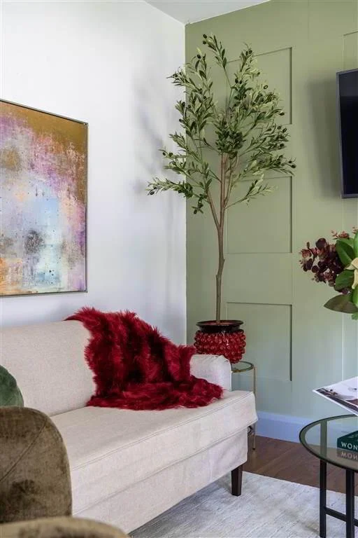 Living room with a beige sofa with a red furry throw, a colorful abstract painting on the white wall, a tall potted plant in a red pot, and part of a glass coffee table with a magazine on it.