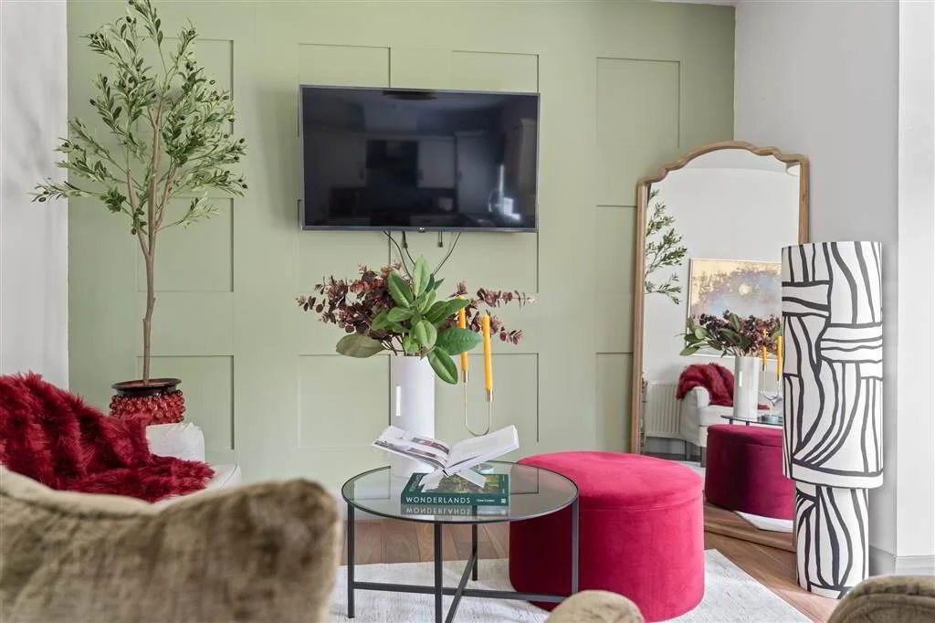 Living room with a wall-mounted flat-screen TV, a large mirror, a potted tree, a red plush ottoman, a glass coffee table with a book and a vase of flowers, a patterned floor lamp, and a sofa with red and beige throws.