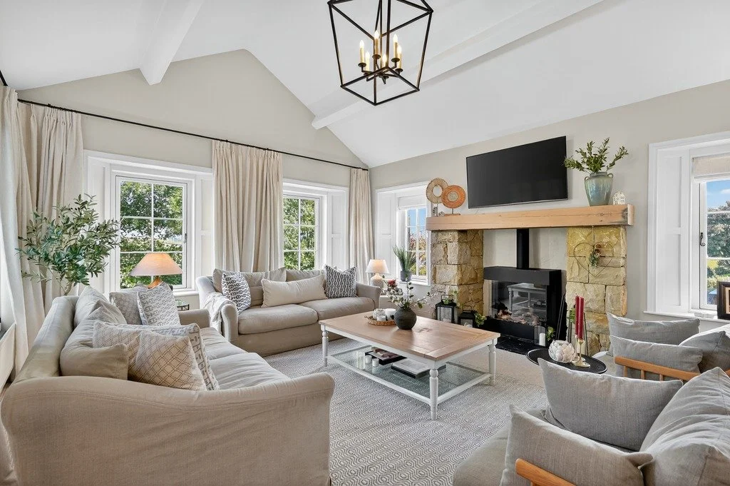 Bright living room with beige sofas, large windows with cream curtains, a stone fireplace with a wooden mantel, a mounted flat-screen TV, and decorative plants and pillows.