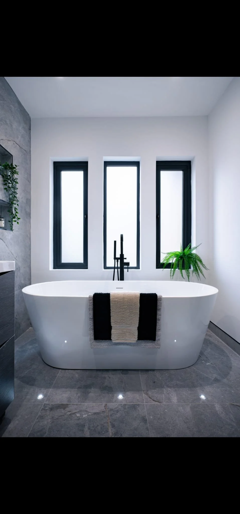 Modern bathroom with a white freestanding bathtub under three tall, narrow windows with black frames, a black faucet, a green plant on the right, and a towel hanging on the front of the tub, with gray tiled flooring.