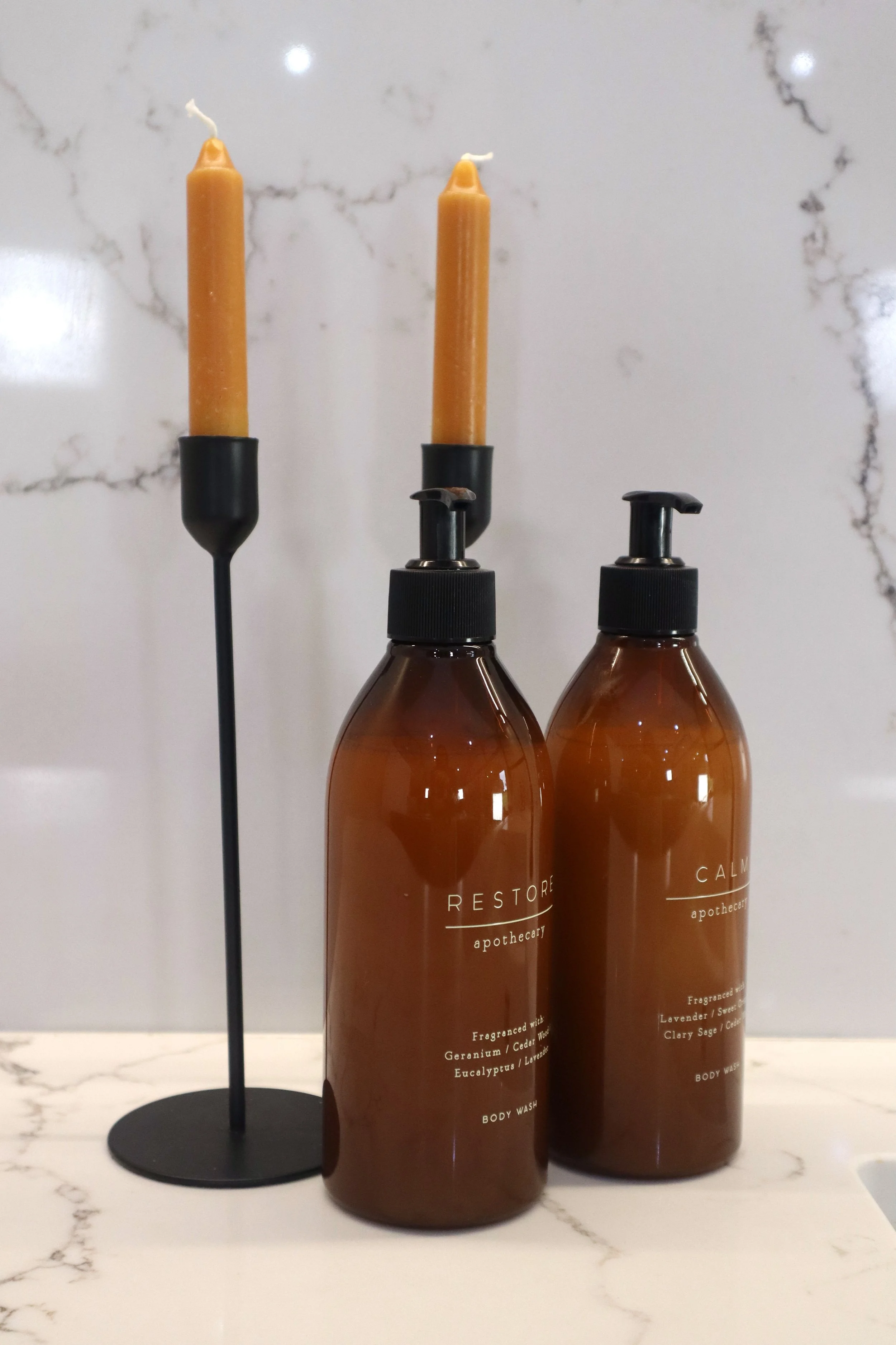 Two amber-colored bottles of body wash labeled 'Restore Apothecary,' standing next to a black candle holder with two unlit yellow candles, on a white marble surface with a white marble wall background.