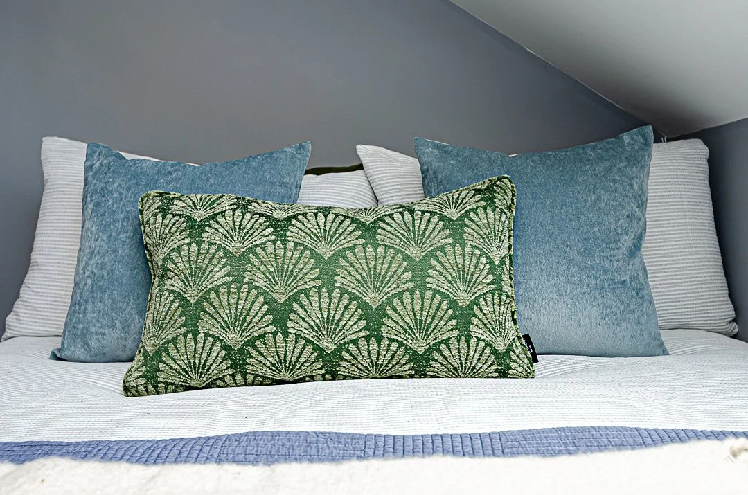 A neatly made bed with a white quilt, a blue blanket at the foot, and multiple decorative pillows, including a prominent green pillow with a fan-shaped pattern and several solid blue and white striped pillows, against a gray wall with a sloped ceilin