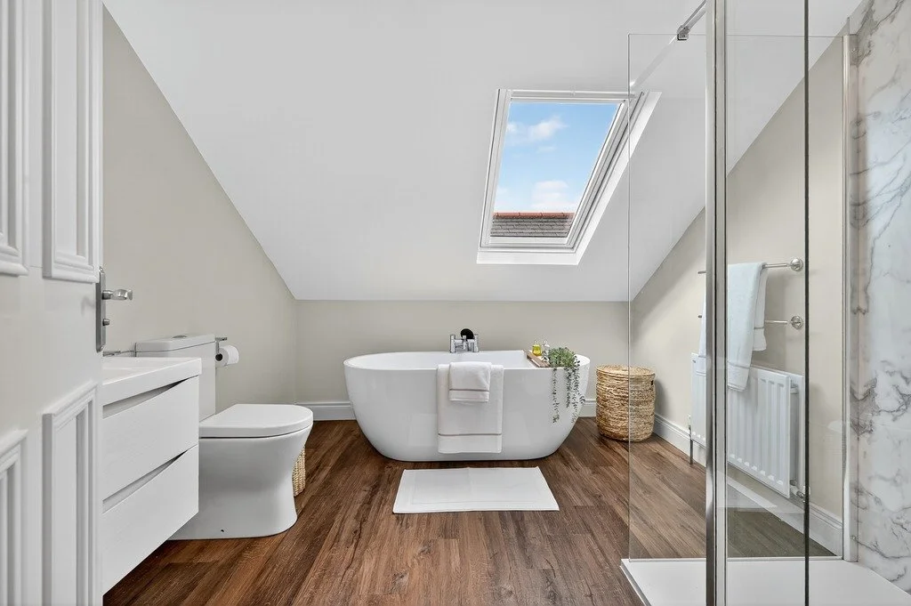 Bright bathroom with a bathtub, toilet, and shower area under a sloped ceiling with a skylight, wood flooring, and neutral-colored walls.