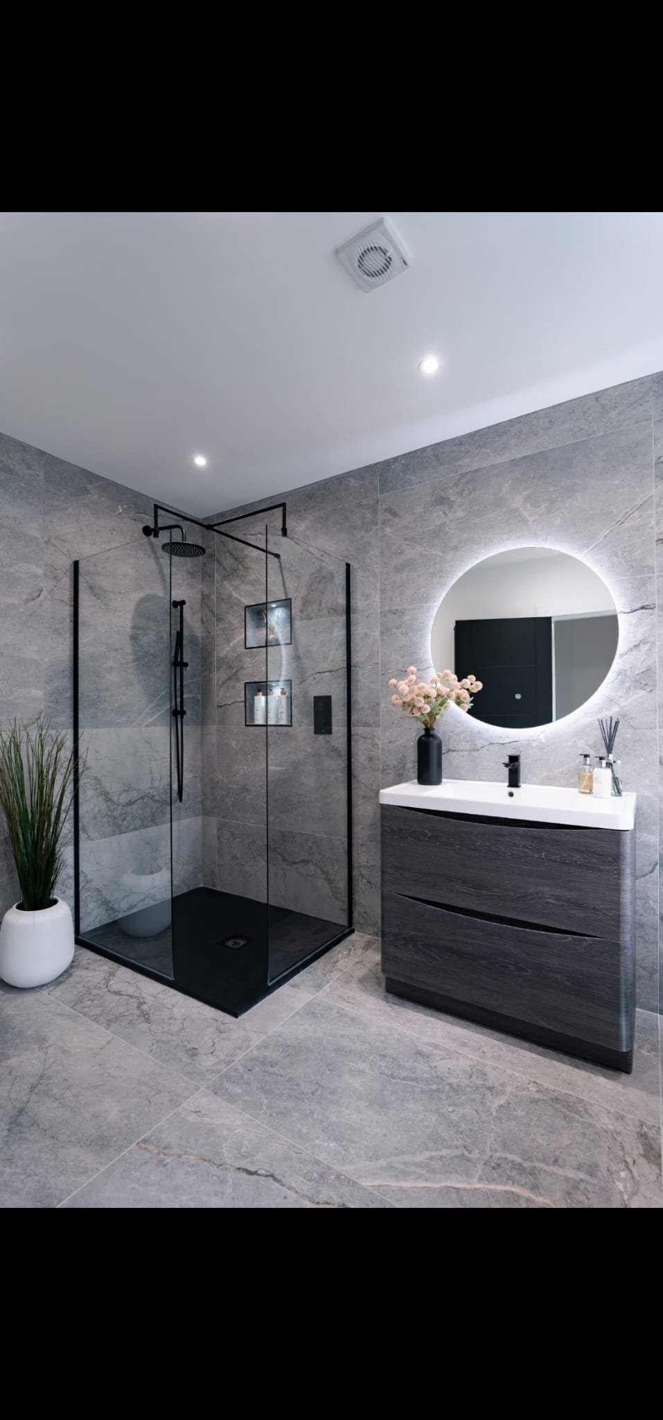 Modern bathroom with gray marble tiles, a glass shower with black fixtures, a round backlit mirror, a dark wood vanity with a white countertop, a black vase with pink flowers, and a tall potted plant.