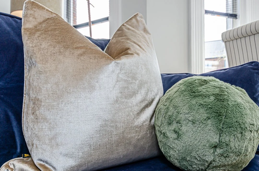 A beige velvet pillow and a round green plush pillow on a dark blue sofa in a bright room with windows.