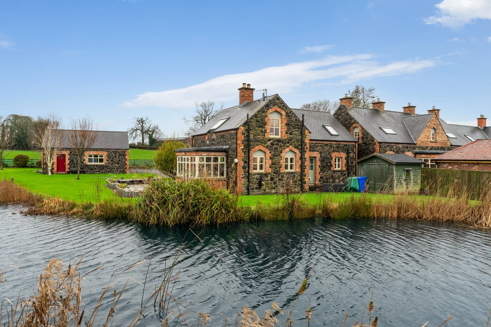 A traditional stone house with a dark slate roof is situated beside a pond, surrounded by green grass, trees, and other houses in a rural landscape under a partly cloudy sky.