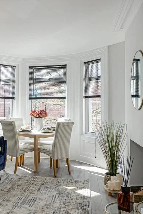 A bright dining area with a round table, four upholstered chairs, a vase of pink flowers, large bay windows with black blinds, a gray rug, and a decorative mirror on the wall.