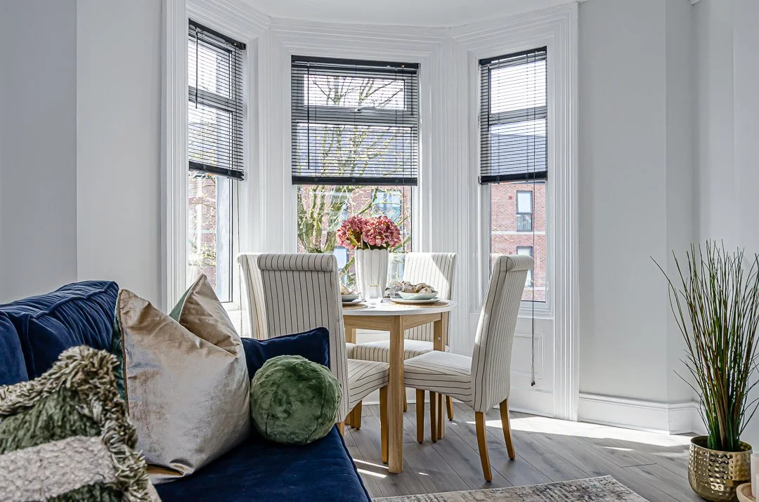 A dining area with a round wooden table set with white dishes and a pink flower centerpiece, surrounded by four upholstered chairs with white and beige striped fabric, located near large bay windows with black blinds in a bright, modern living space.