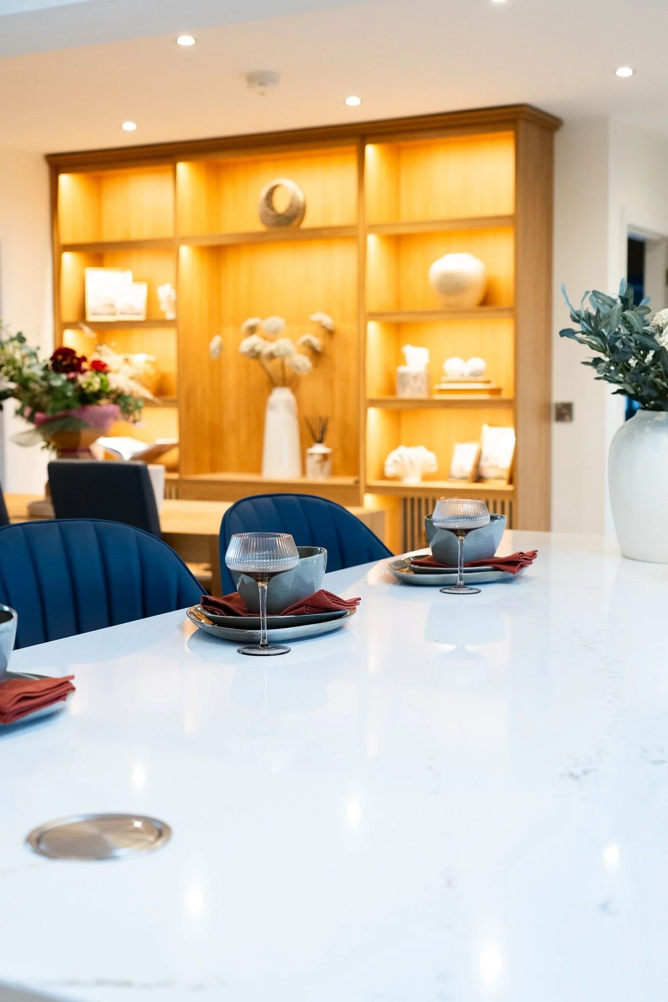 A modern dining room with a white countertop table set with bowls, glasses, and napkins, and a wooden shelving unit with decorative items and flowers in the background.