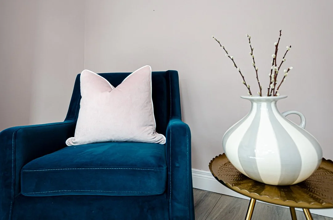 A blue upholstered armchair with a light pink velvet pillow in a room with a beige wall. Next to the chair is a small gold side table with a white ceramic vase holding brown branches with small white buds.